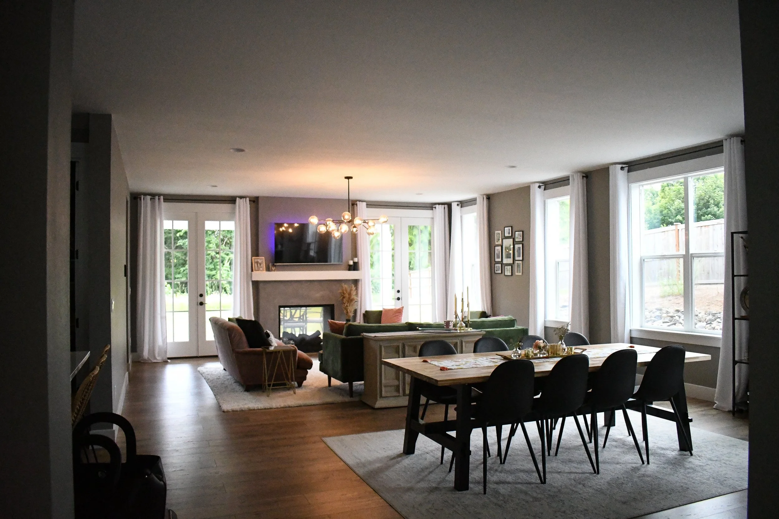Bright living room and dining area with large windows, white curtains, a fireplace, wall-mounted TV, a chandelier, and a wooden dining table with black chairs.
