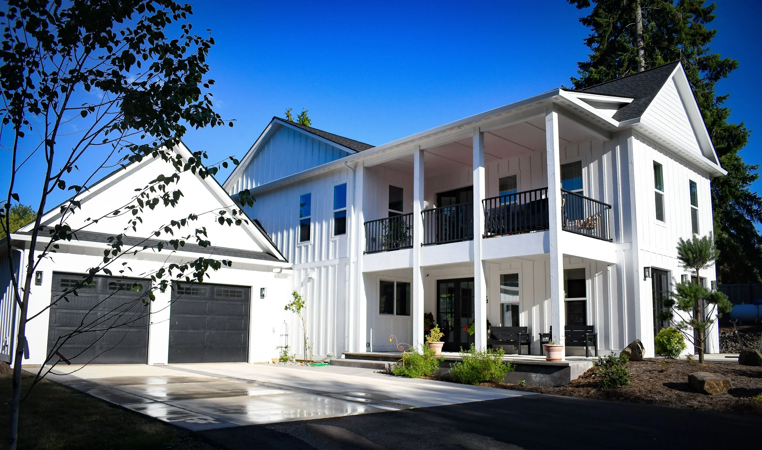 A modern two-story white house with a black roof, large windows, and a balcony with black railing. The front yard has a driveway, small plants, and benches.