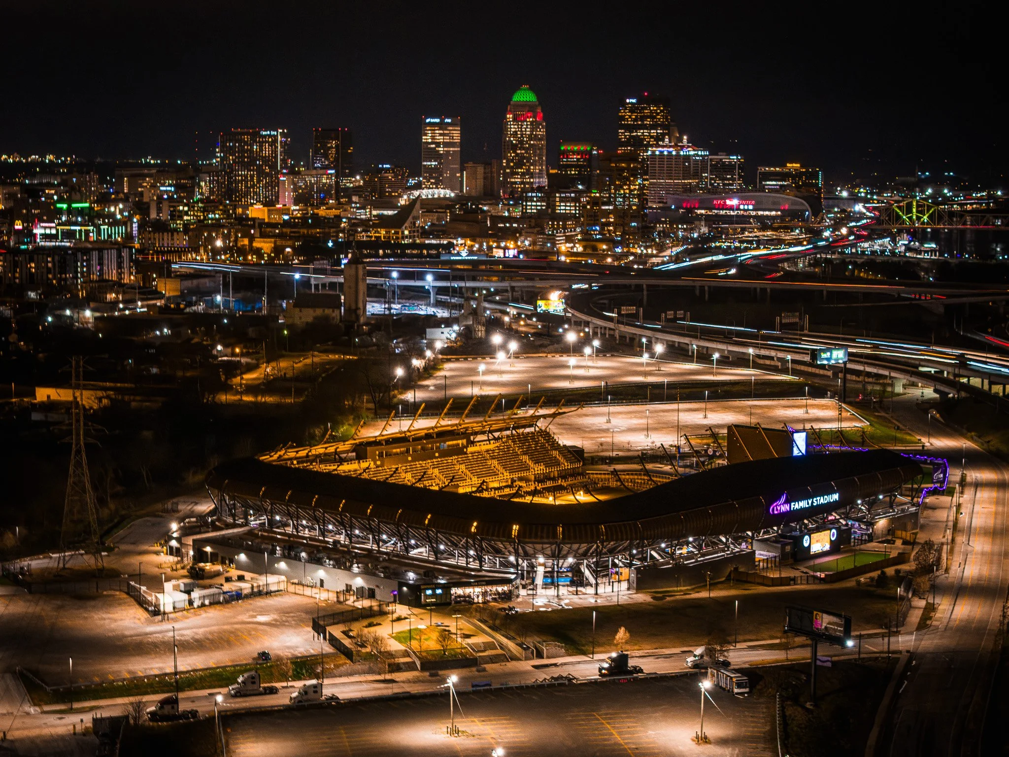 An aerial view of a residential construction site with multiple buildings in various stages of completion, surrounded by open fields and nearby neighborhoods