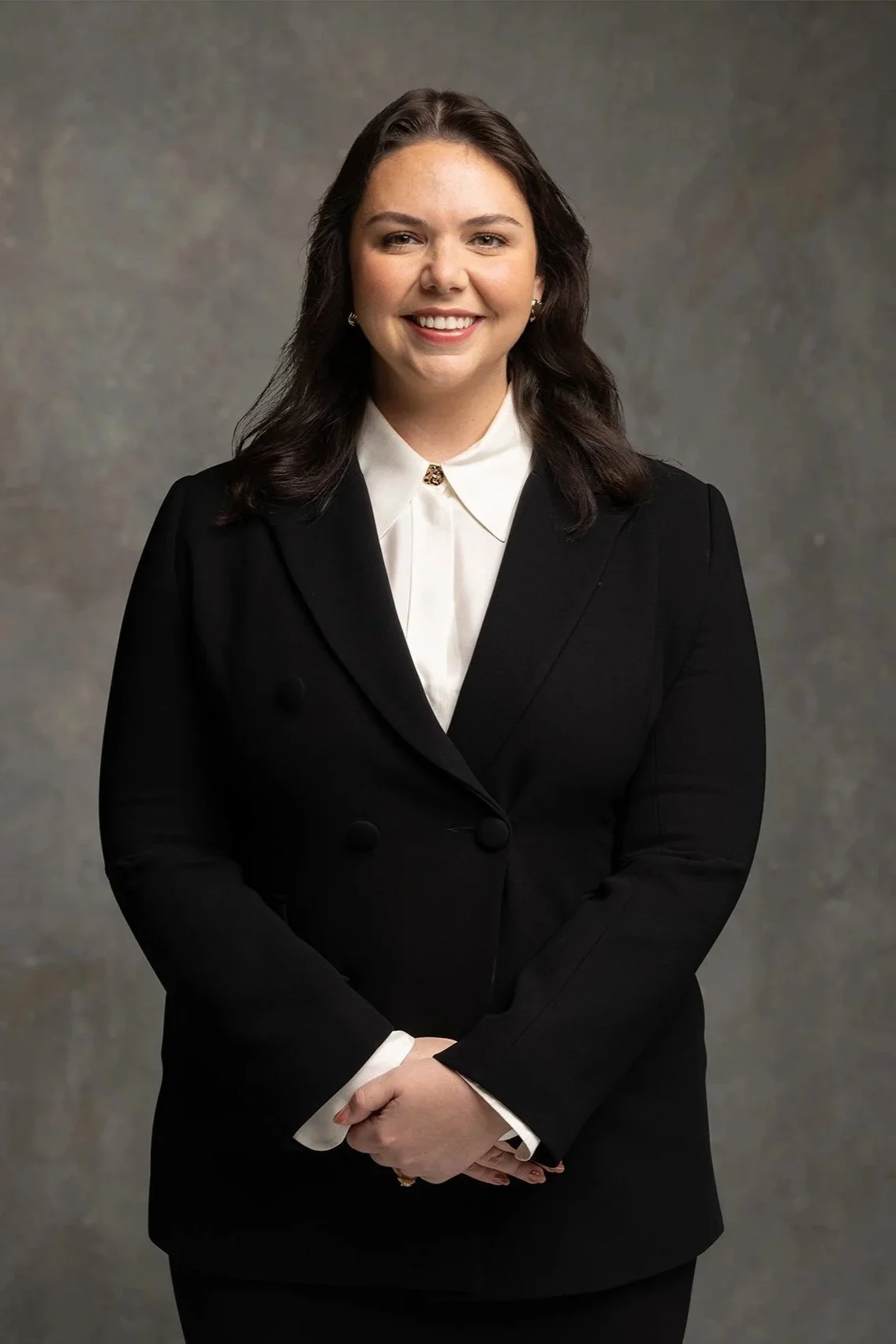 A woman with dark brown hair, smiling, dressed in a black blazer over a white collared shirt, standing against a gray textured background.
