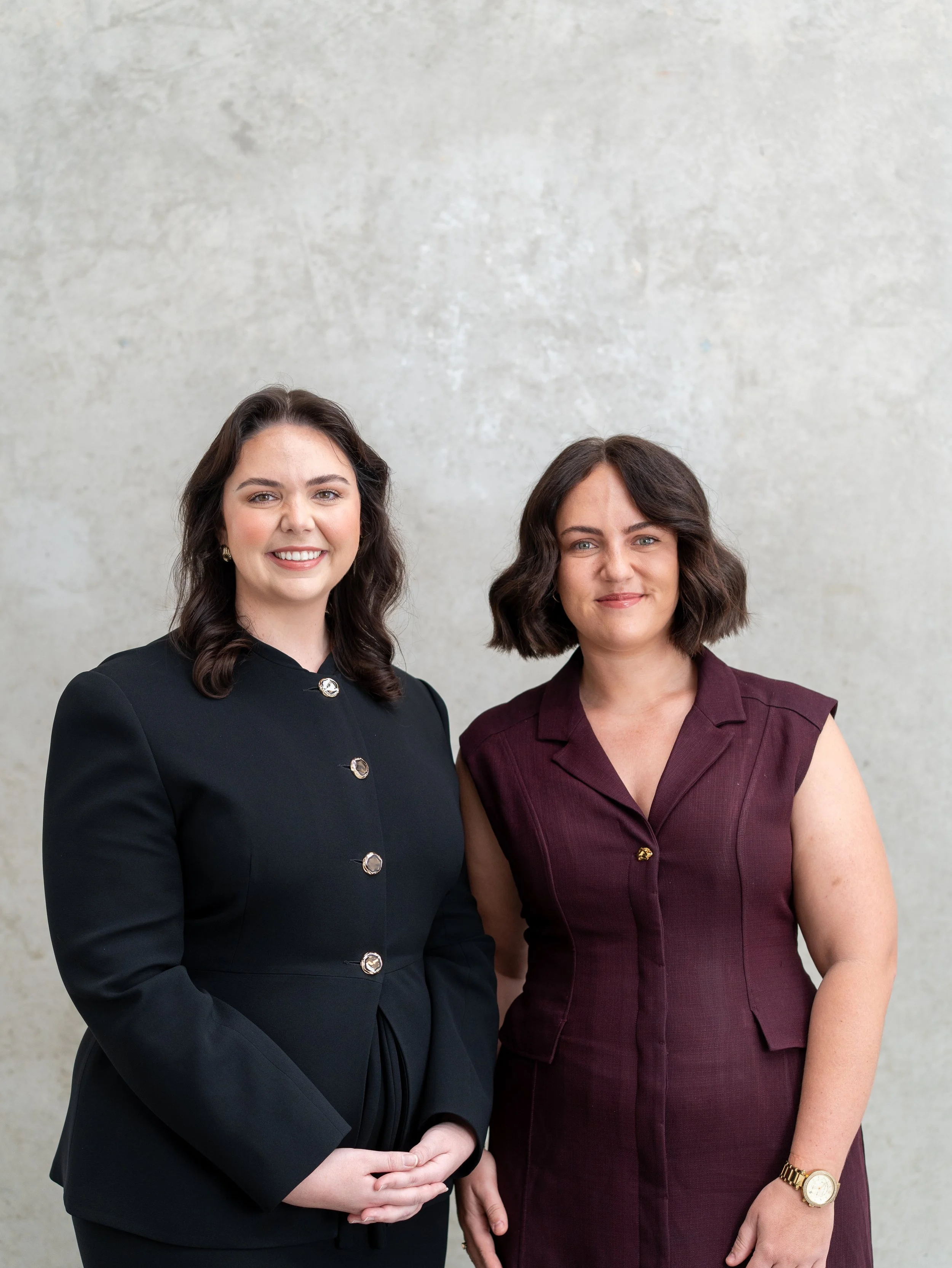 Two women standing side by side, wearing business attire, and smiling at the camera against a plain light-colored wall.