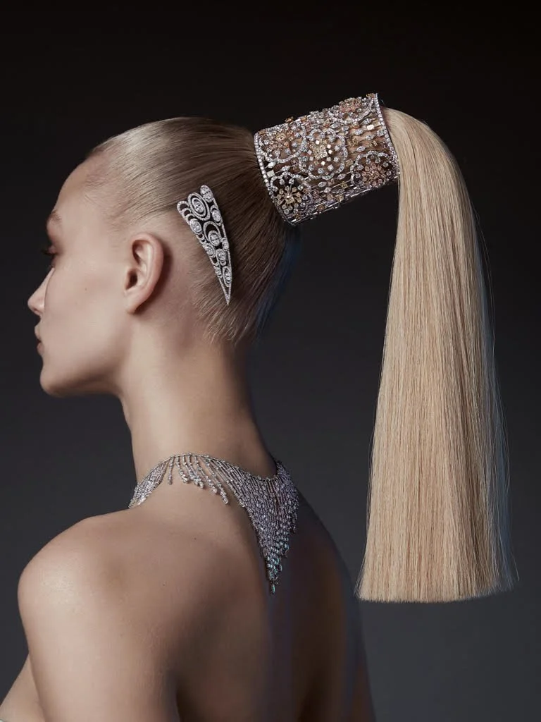 A woman with sleek, blonde hair styled in a high ponytail decorated with rhinestone jewelry and a large, sparkly hairpiece. She is wearing an elaborate, sparkling necklace and a matching earring, against a dark background.