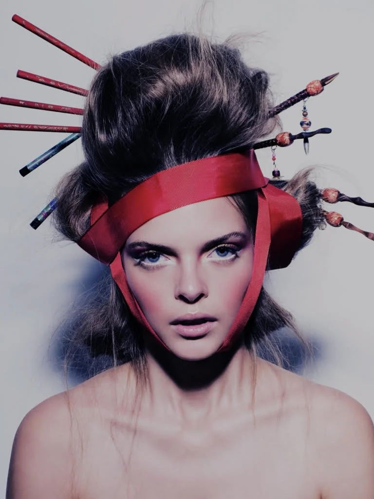 A woman with elaborate hair styled in a high, voluminous updo, adorned with multiple decorative hairpins. She wears a red ribbon around her head and face, with the ribbon tied under her chin. She has bright blue eyes, bold makeup, and a neutral expression.