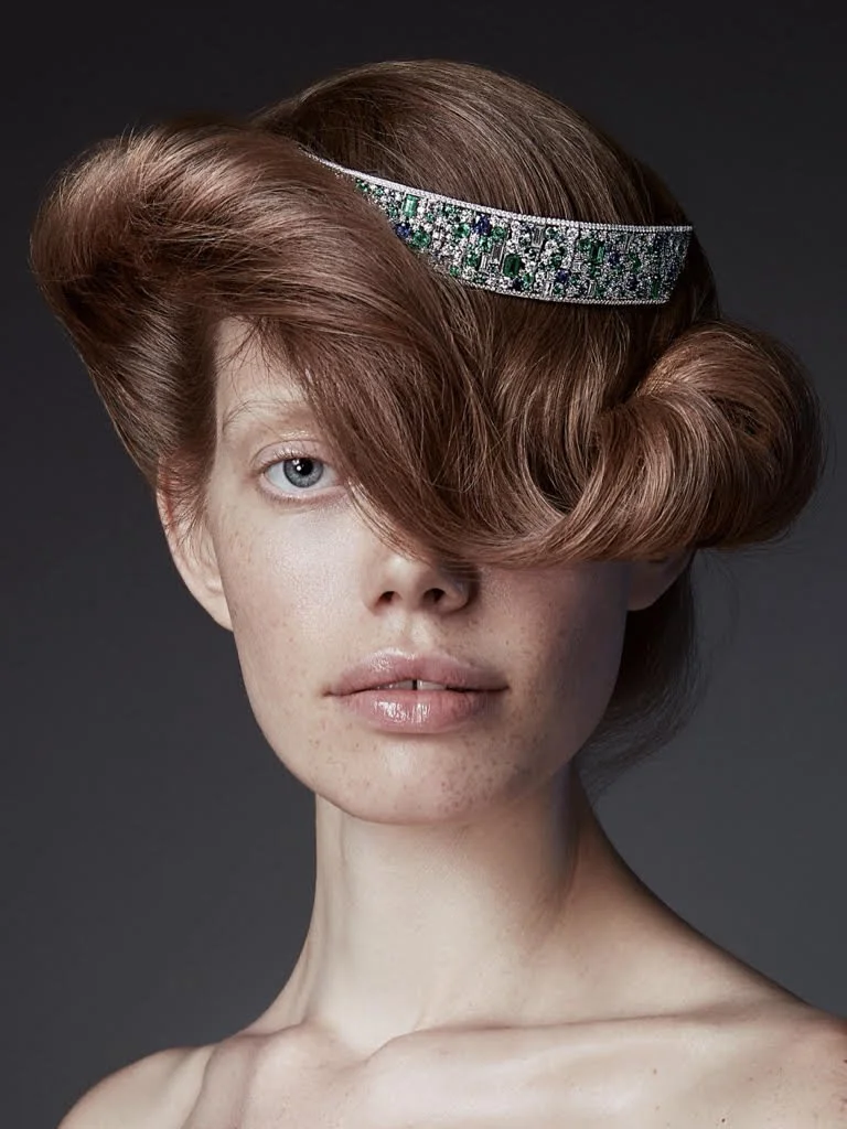 A woman with red hair styled in a vintage updo, wearing a bejeweled headband, with one eye partially covered by her hair, and looking directly at the camera.