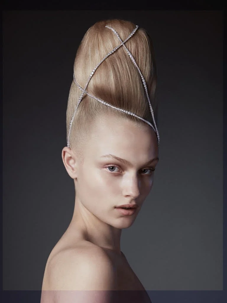 A woman with an avant-garde hairstyle featuring a voluminous, tall updo with a golden hue, decorated with silver, crisscrossing jewelry accents against a dark background.