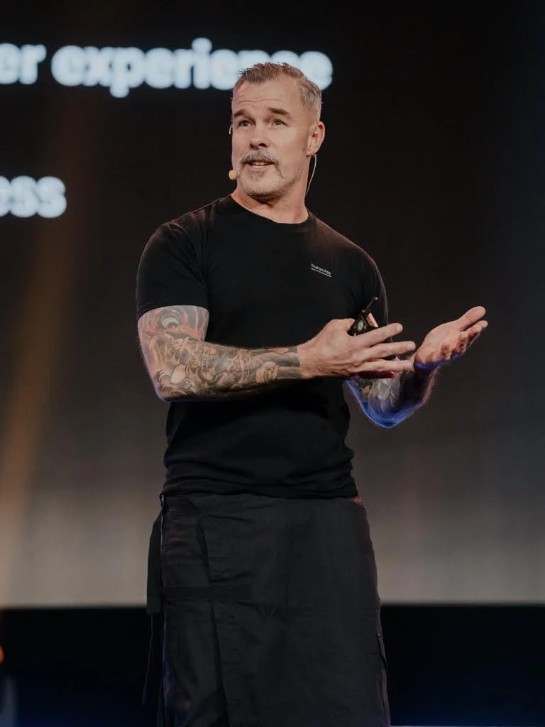 A man with tattoos on his arms is speaking on stage, holding a microphone and a remote control, wearing a black t-shirt and black pants, with a presentation slide visible in the background.