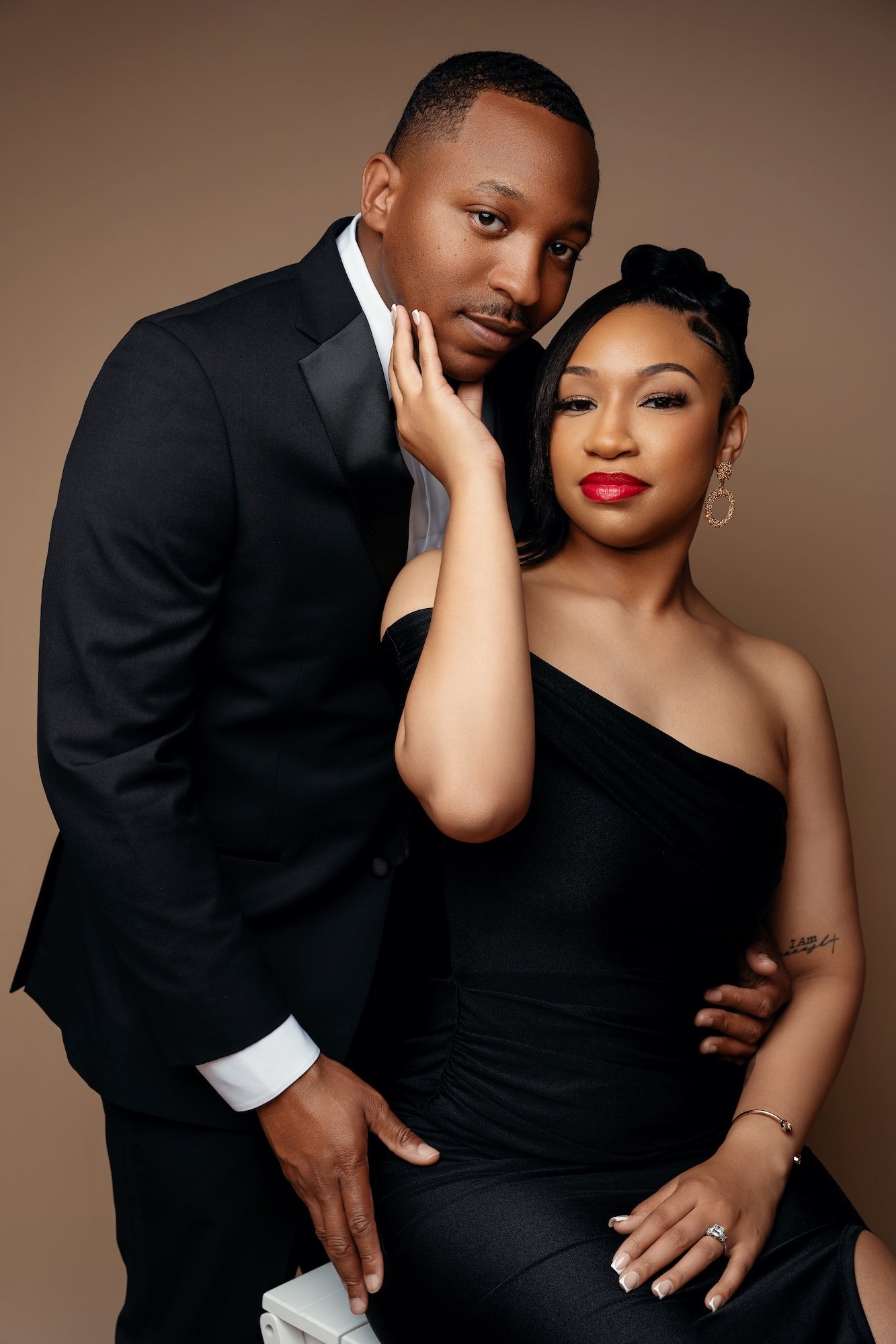 Intimate engagement portrait, woman in black one-shoulder dress touching man's face, brown backdrop, romantic studio photography Chicago