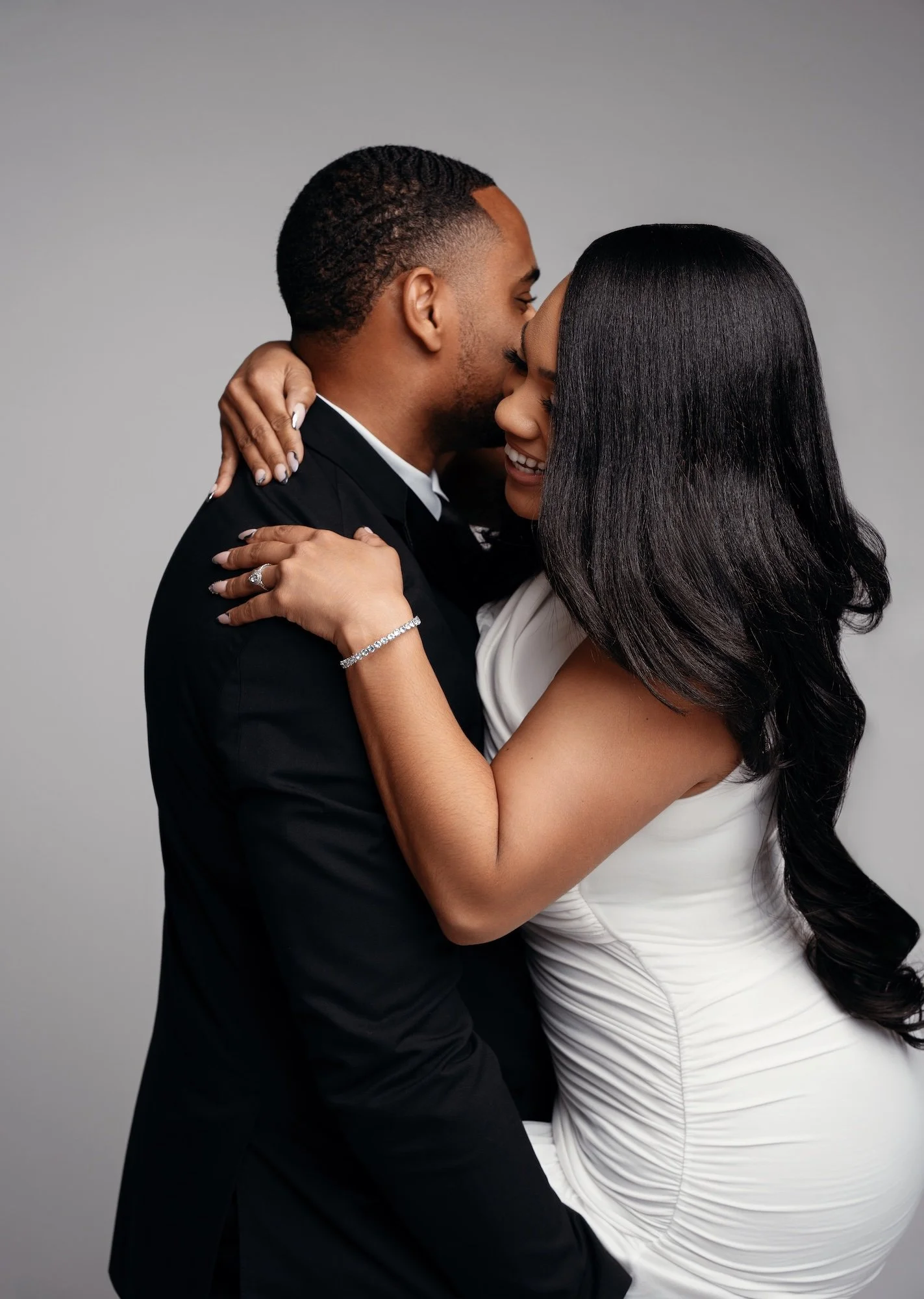 Intimate engagement embrace, man in black suit holding woman in white dress close, gray backdrop, romantic studio portrait Chicago
