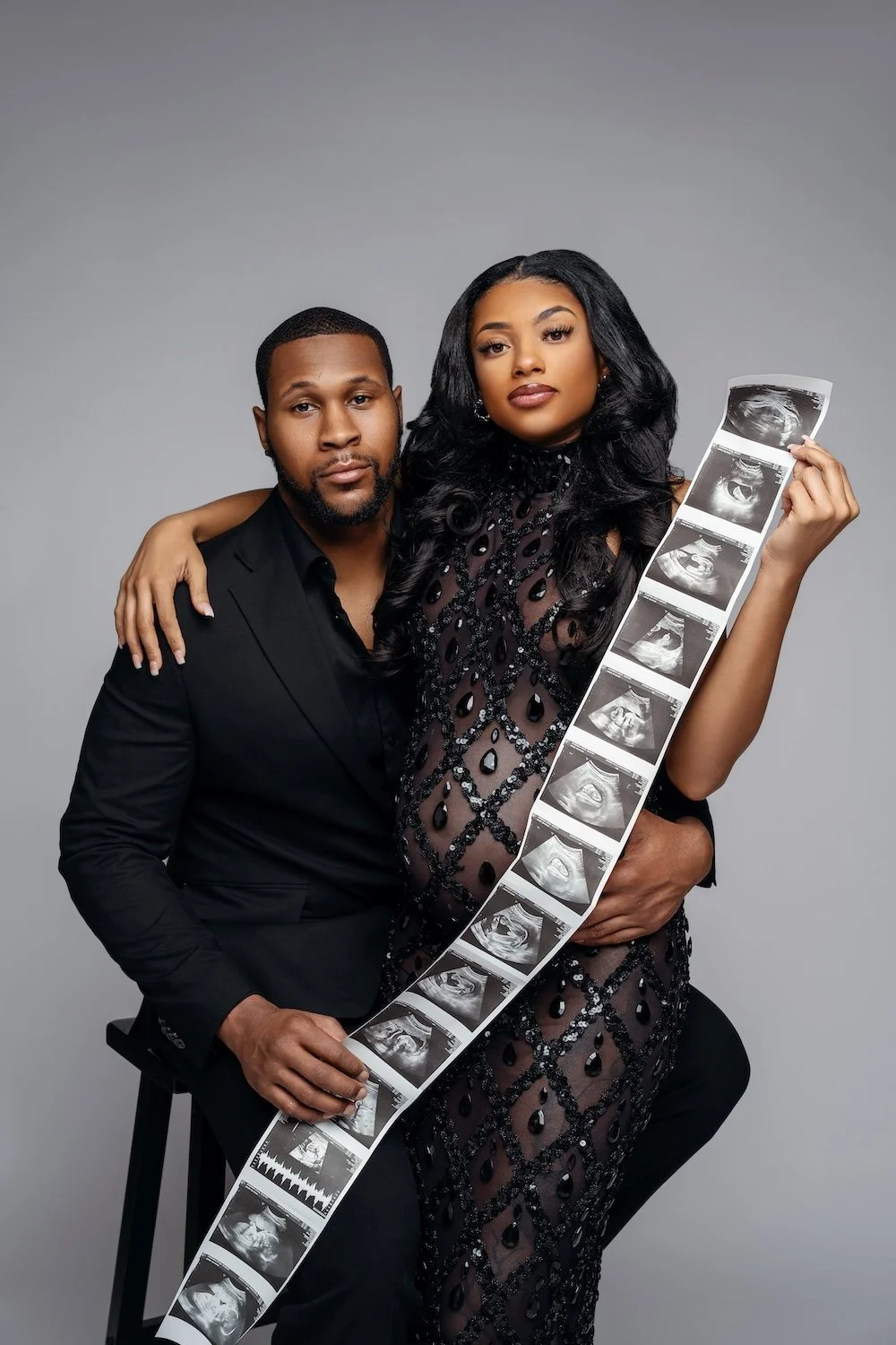 Stylish expecting couple in black attire with ultrasound photo strip, maternity photoshoot