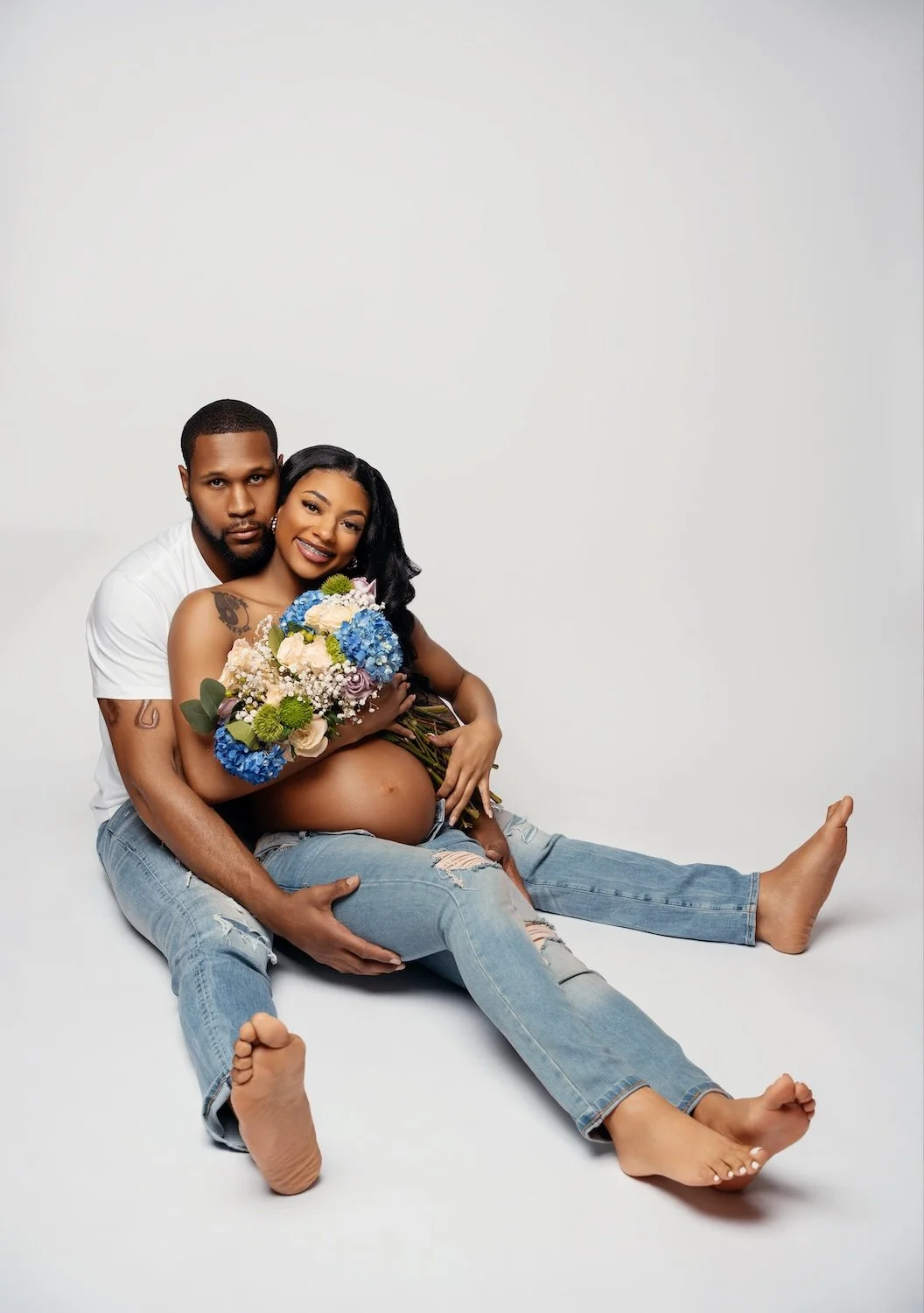 Happy pregnant couple posing barefoot on the floor, woman cradling a blue hydrangea bouquet on her belly while partner embraces her from behind