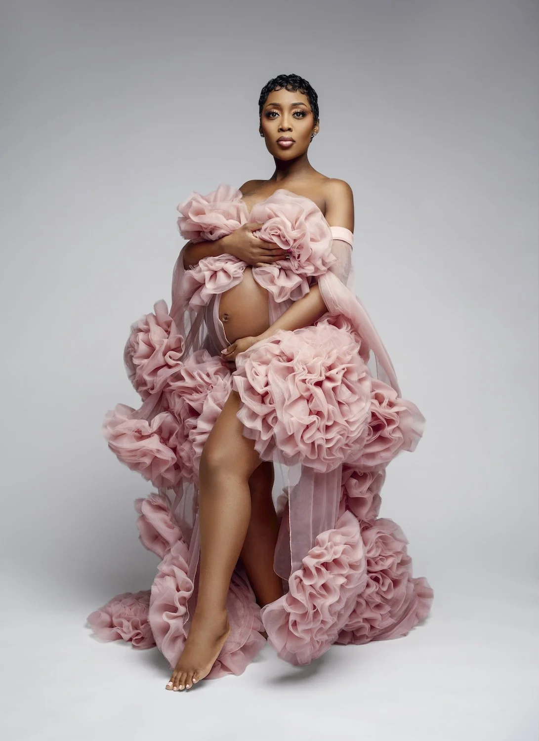 An elegant maternity portrait of a woman standing barefoot. She is draped in a large, sculptural pink tulle fabric with floral-like ruffles, strategically styled to reveal her baby bump and one leg