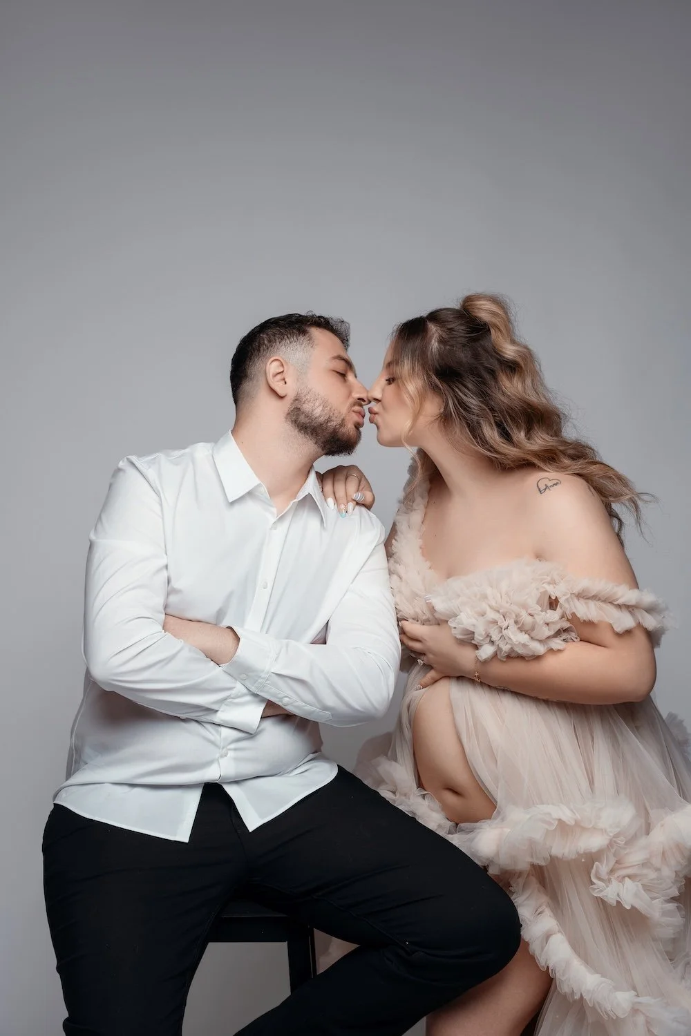 An affectionate studio photograph of an expecting couple against a plain grey background