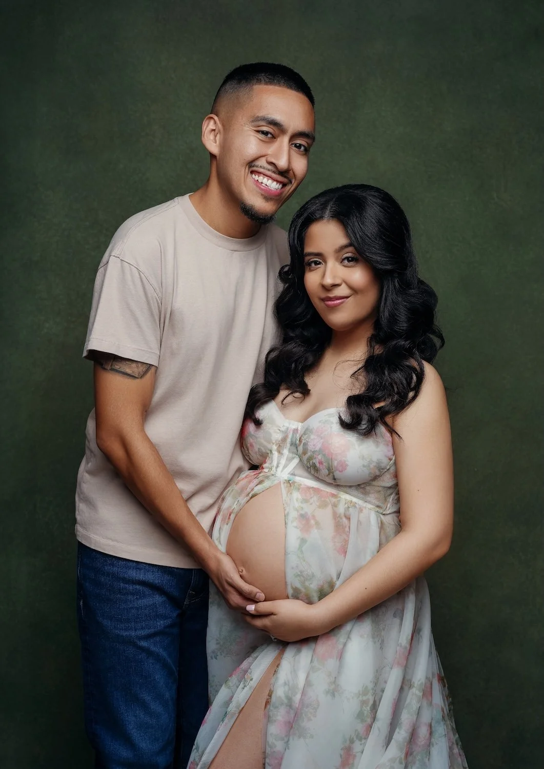 Maternity photo shoot portrait of a smiling couple. He wears a beige tee and she a sheer floral gown, both holding her baby bump against a green backdrop.