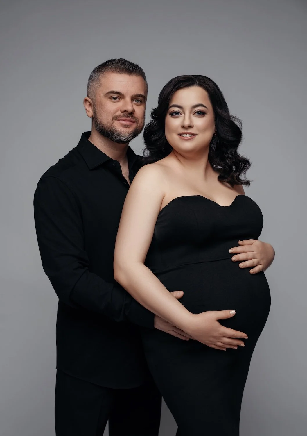 Maternity photo shoot of a smiling couple in elegant black attire against a grey studio background. The man stands behind the pregnant woman, both gently holding her baby bump.