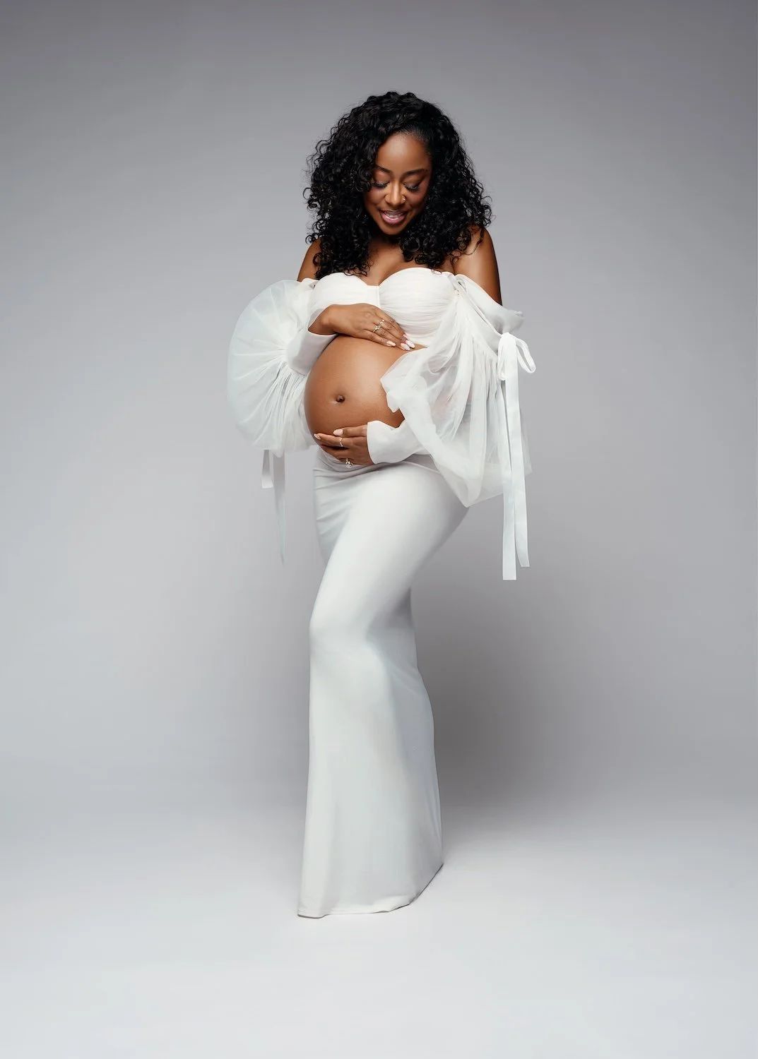  Elegant studio maternity portrait of a pregnant woman with curly hair wearing a white off-the-shoulder gown with sheer puffy sleeves and a fitted mermaid skirt, smiling as she gently cradles her baby bump against a soft grey backdrop. 