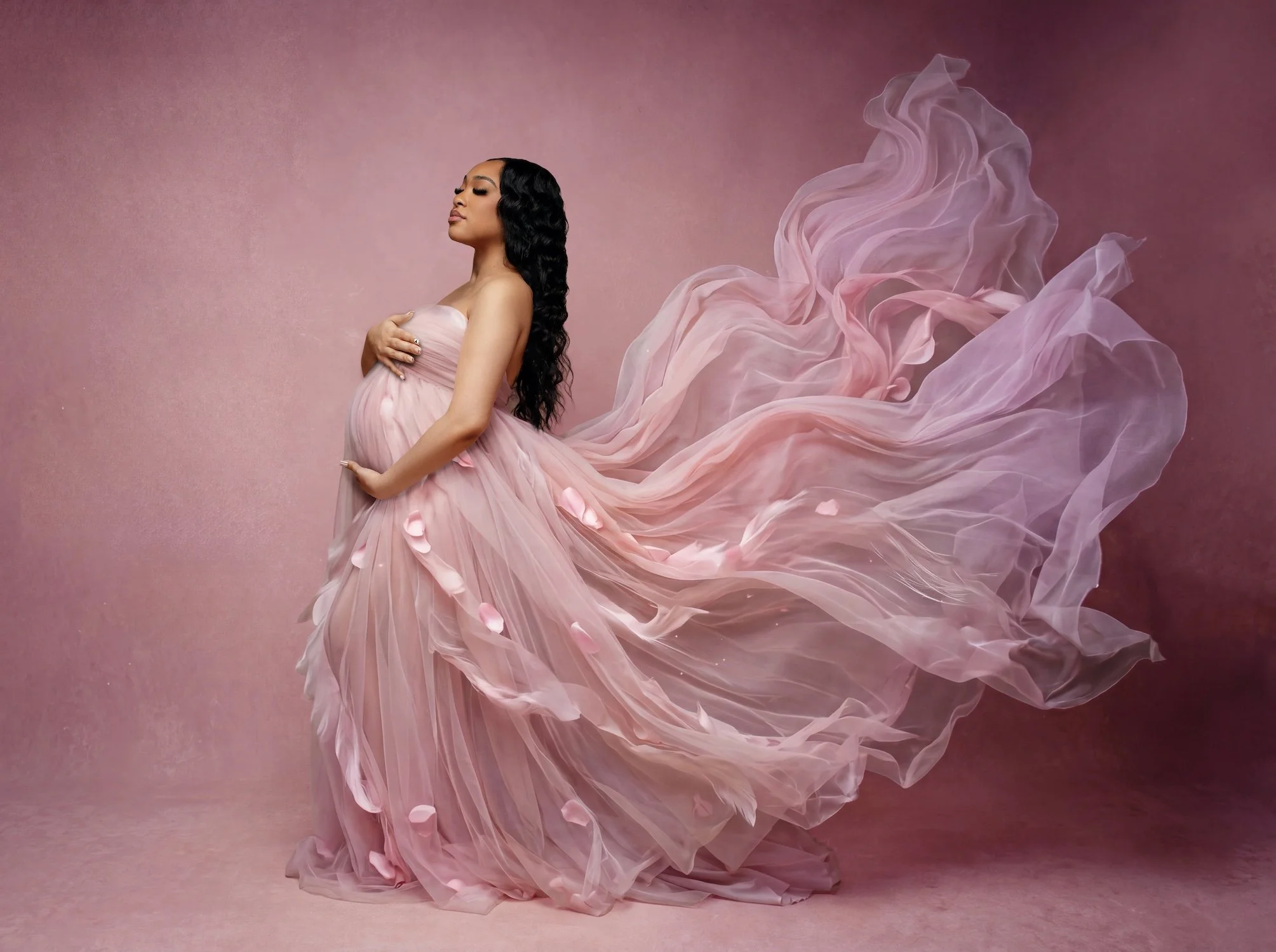  Ethereal maternity portrait of a pregnant woman in profile wearing a flowing pink chiffon gown adorned with scattered petals, with the fabric billowing behind her like wings against a soft mauve studio backdrop. 