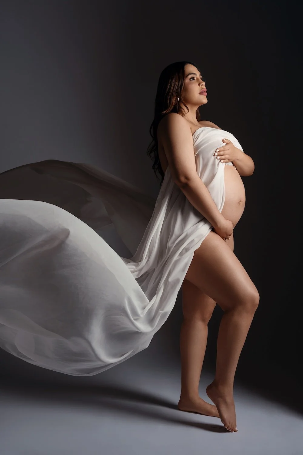  Pregnant woman in flowing white fabric, posing in a dramatic dark studio maternity portrait. 