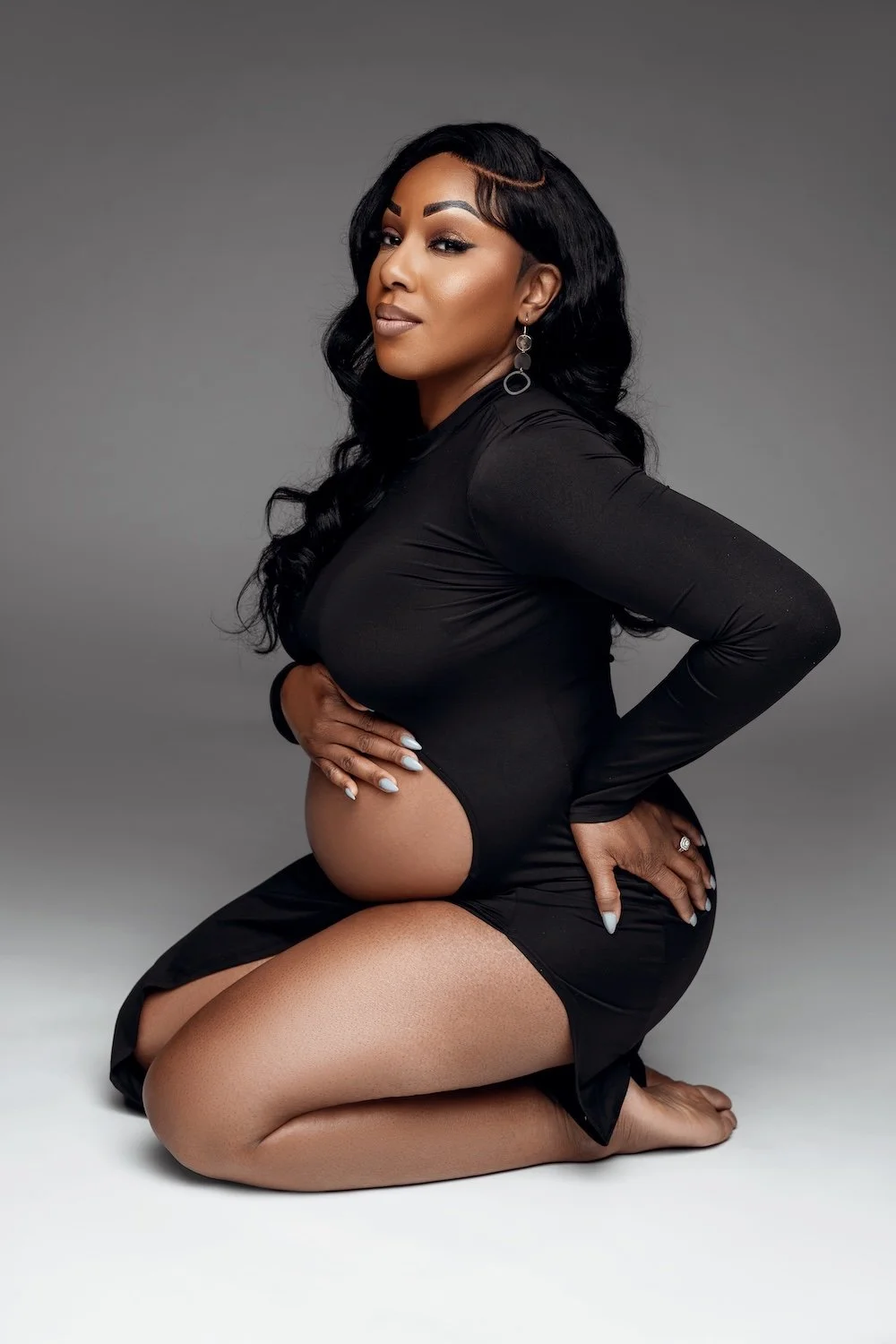  Pregnant woman kneeling in a black dress, posing in a studio maternity portrait with a soft gray background. 