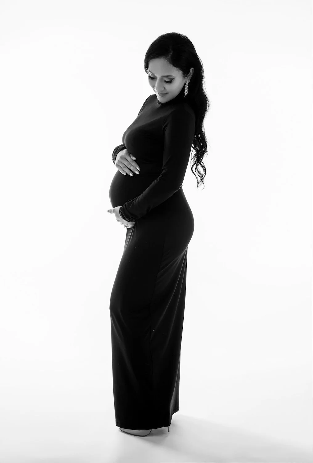  Black and white maternity photography Chicago - pregnant woman in a long fitted black gown, hands cradling her belly, soft light against a white studio backdrop. 