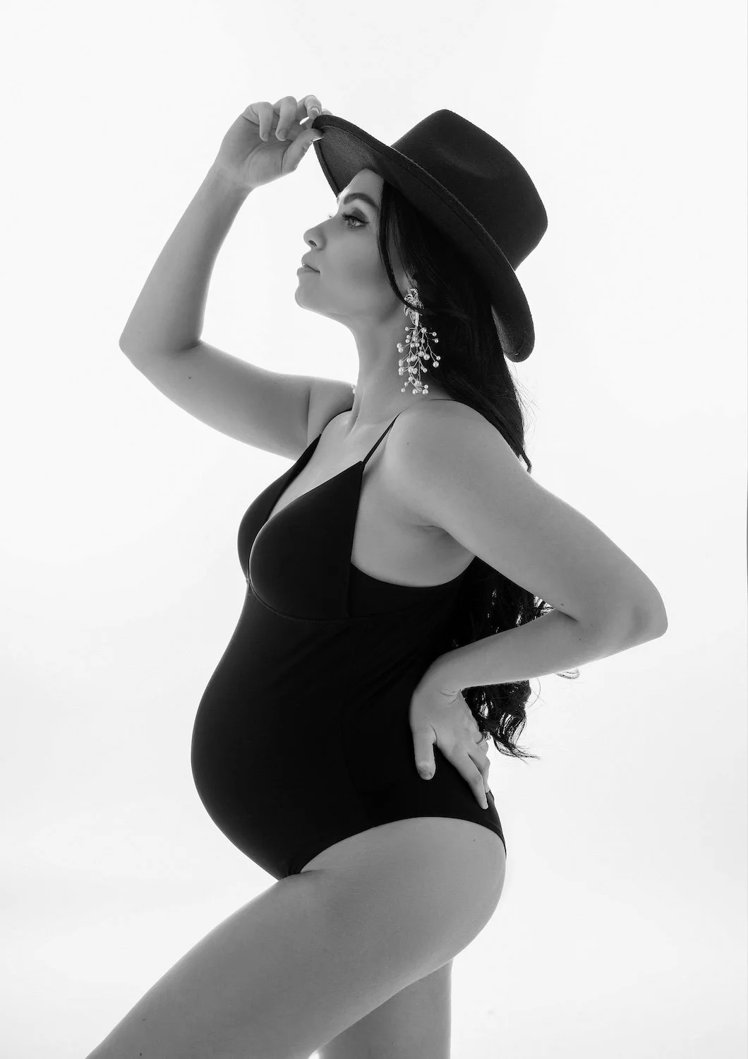  Black and white maternity portrait in our Chicago studio - pregnant woman in a black bodysuit and wide-brim hat, side profile against a clean white backdrop. 