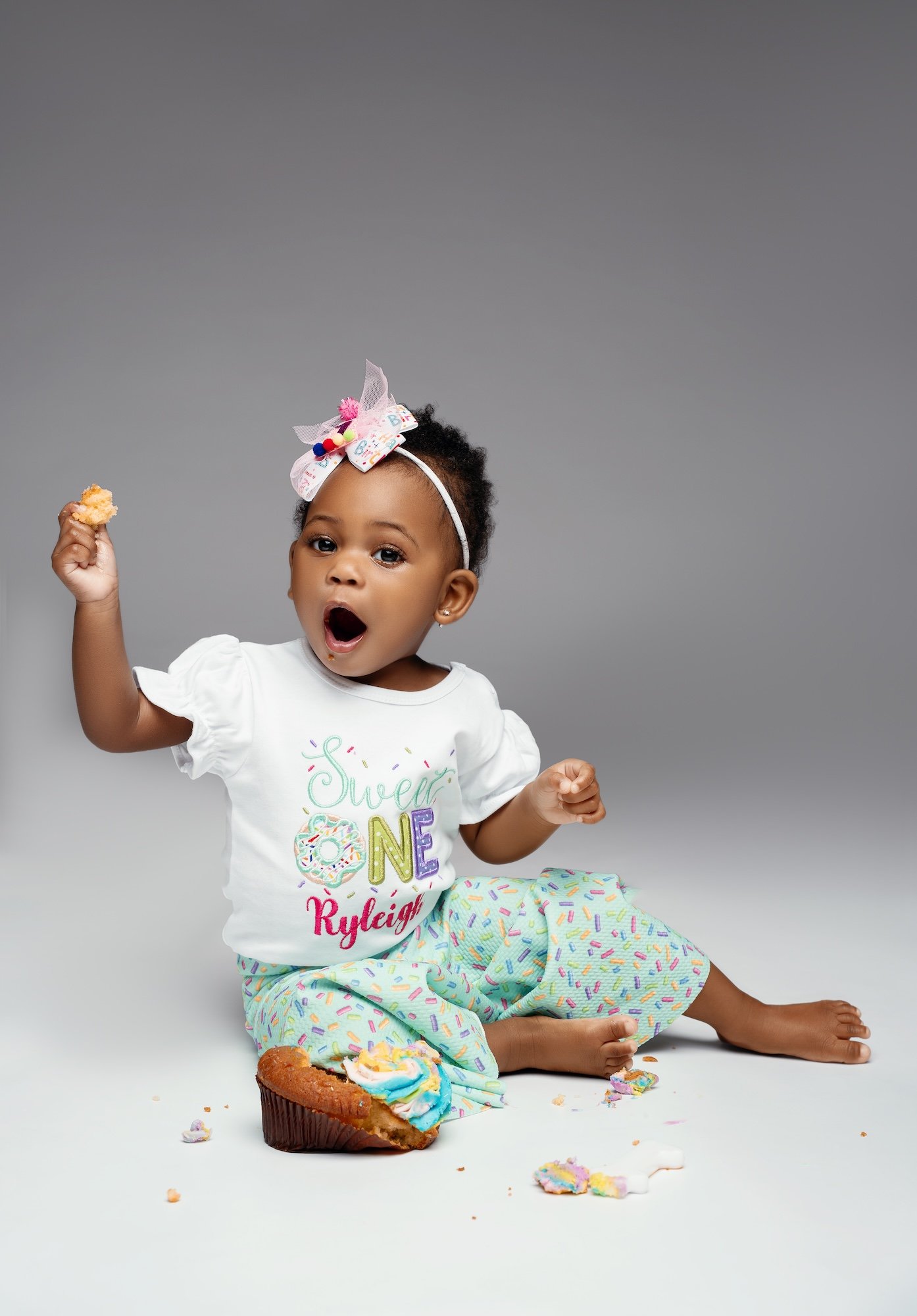  Baby girl in Sweet One outfit holding cupcake during studio cake smash — joyful first birthday photo session 