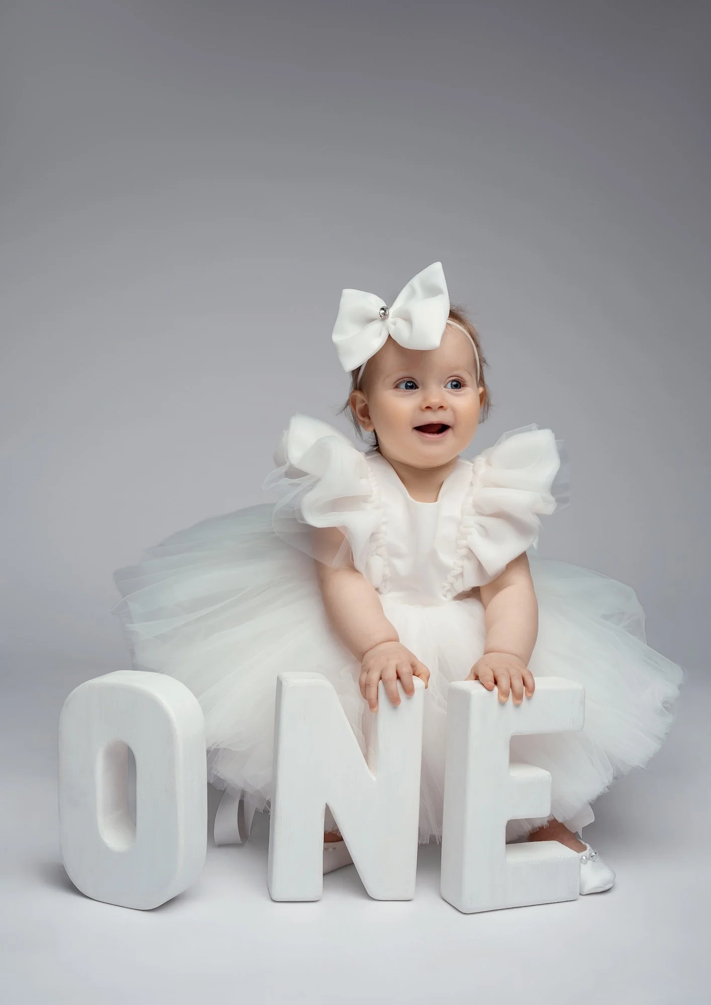  Smiling baby girl in white dress with bow holding large ONE letters — studio first birthday portrait 