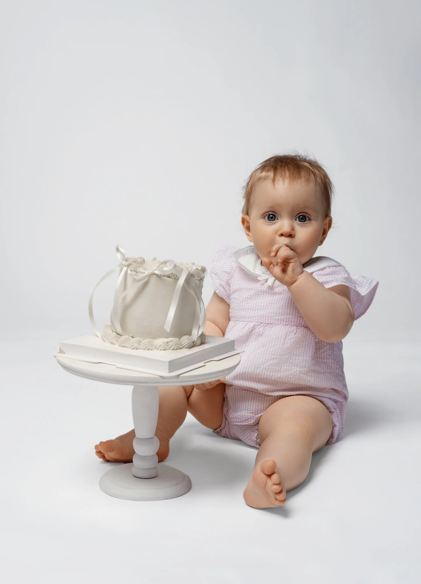  Baby girl in pink outfit sitting next to white cake on stand — studio cake smash session for first birthday 