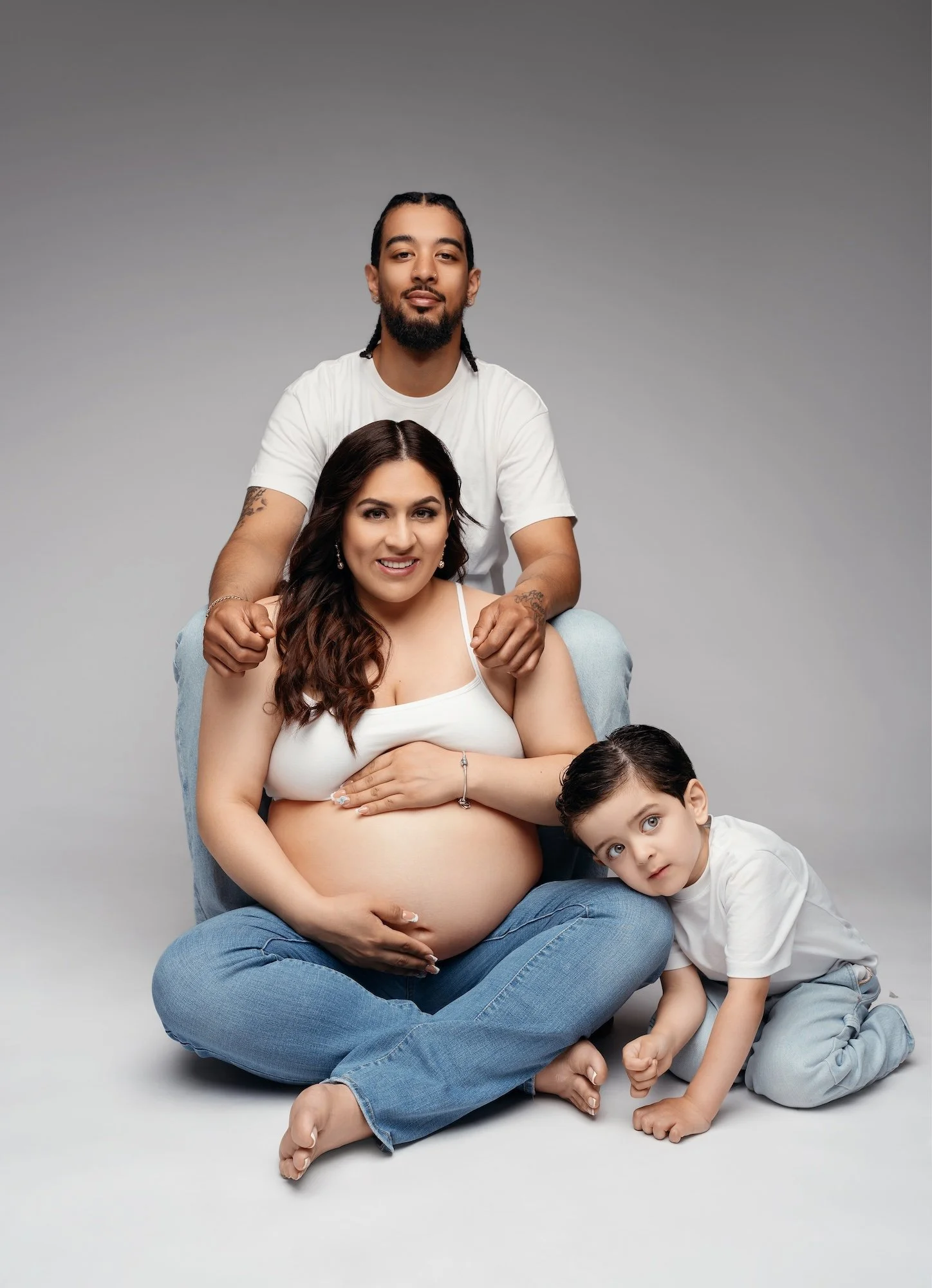  Studio maternity portrait of a family with pregnant mother, father, and young son wearing white tops and jeans on grey background 