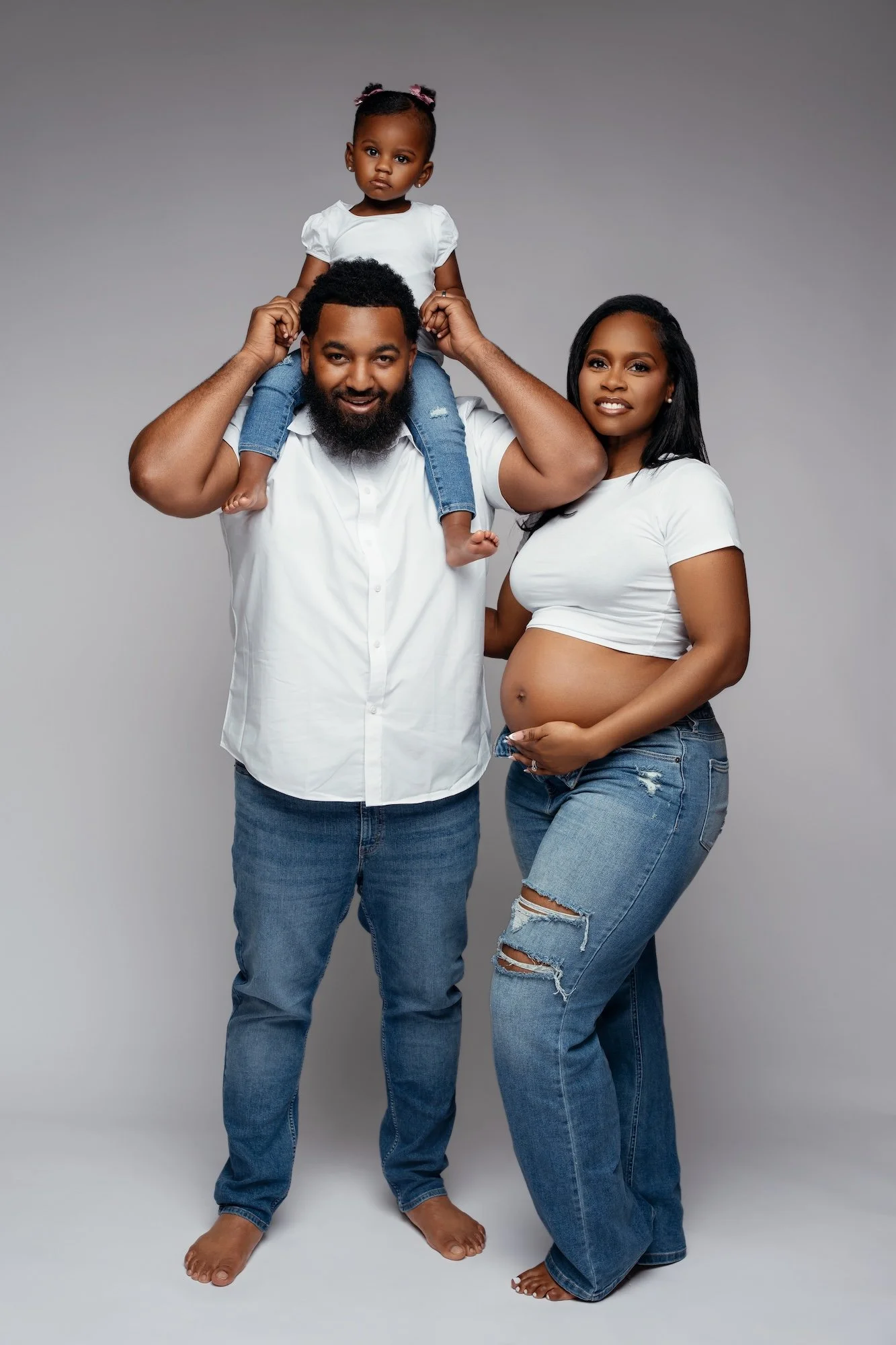  Maternity family portrait with father carrying daughter on shoulders and pregnant mother beside them, grey studio 