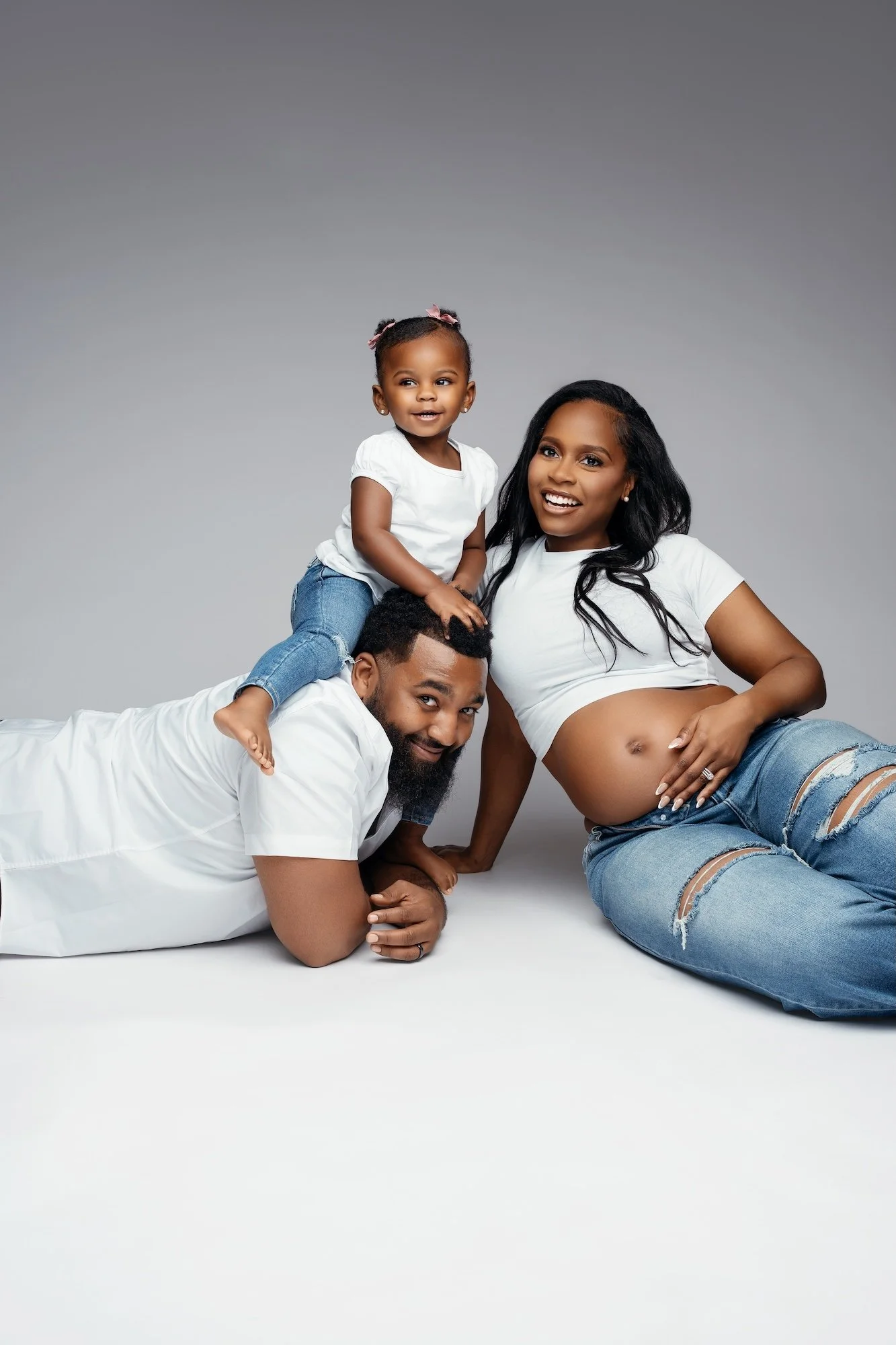  Happy family lying on studio floor with toddler sitting on father's back and pregnant mother resting nearby 
