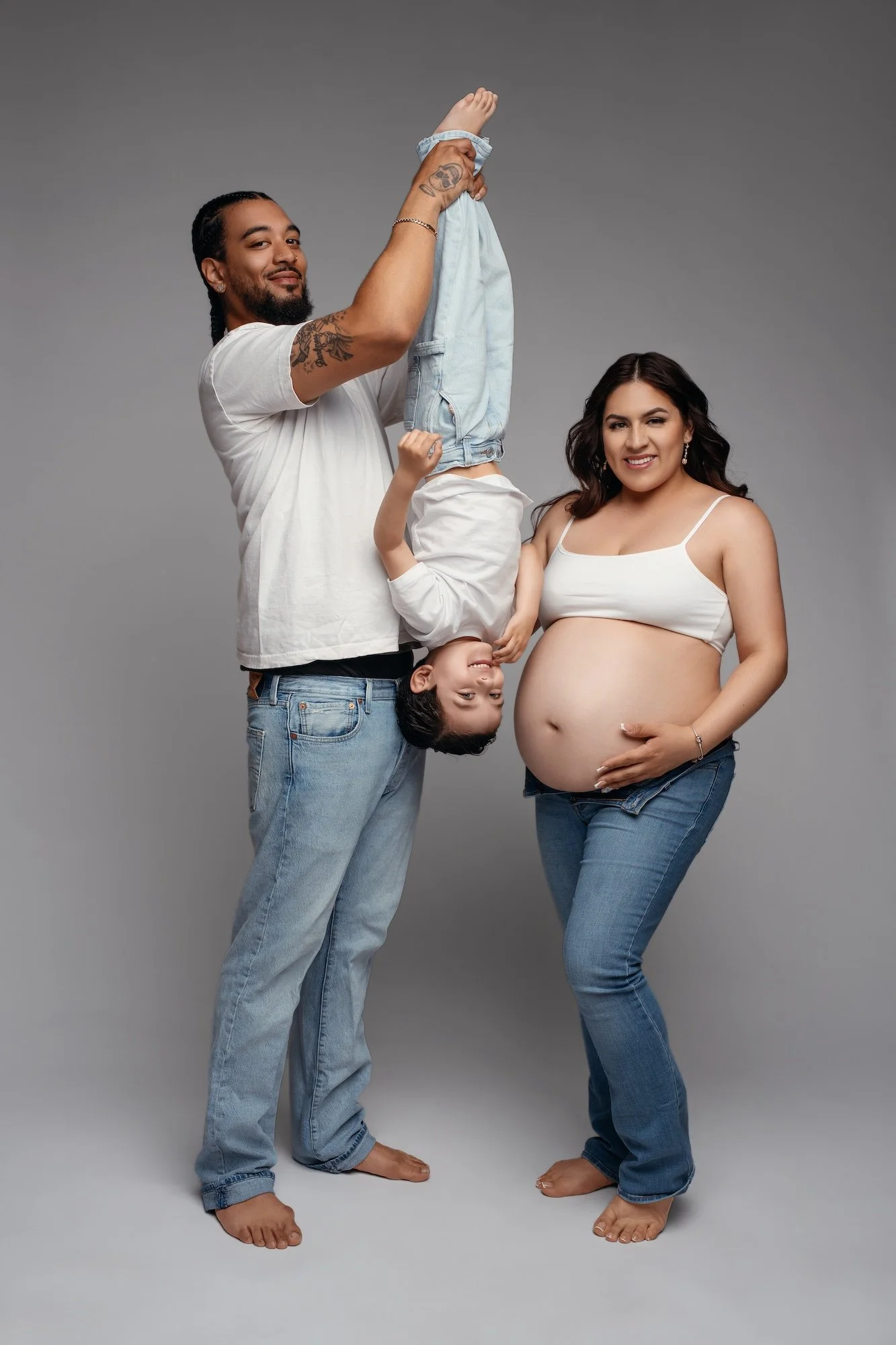  Family maternity photo with father playfully holding toddler upside down next to pregnant mother in studio 