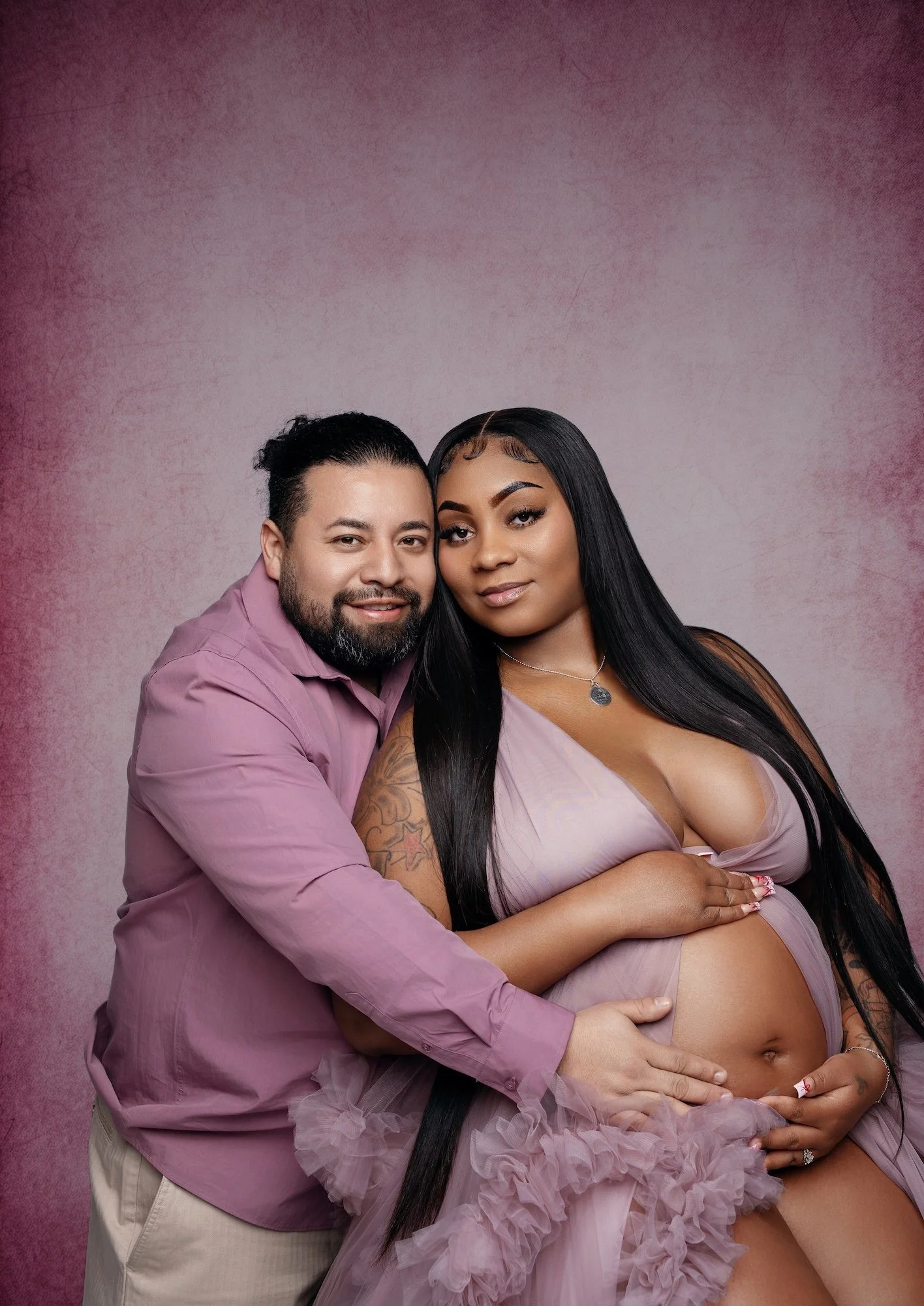 A romantic studio maternity portrait of a smiling pregnant woman with long black hair in a flowing lavender dress, embracing her partner who is wearing a matching purple shirt. They stand close together with his hands gently resting on her pregnant b
