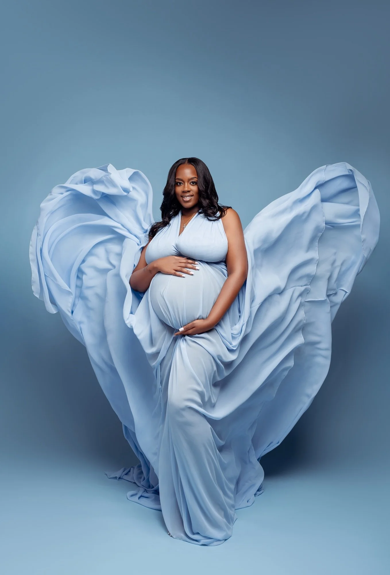  Beautiful pregnant woman in a flowing light blue maternity gown with dramatic fabric wings, hands gently on her belly, studio maternity photography in Chicago by Filaneyka 