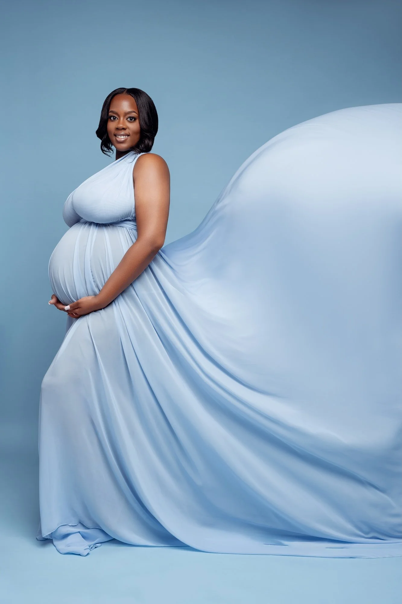  Elegant side profile of a pregnant woman in a luxurious light blue maternity dress with long flowing train, smiling at camera, Chicago maternity studio session by Filaneyka 