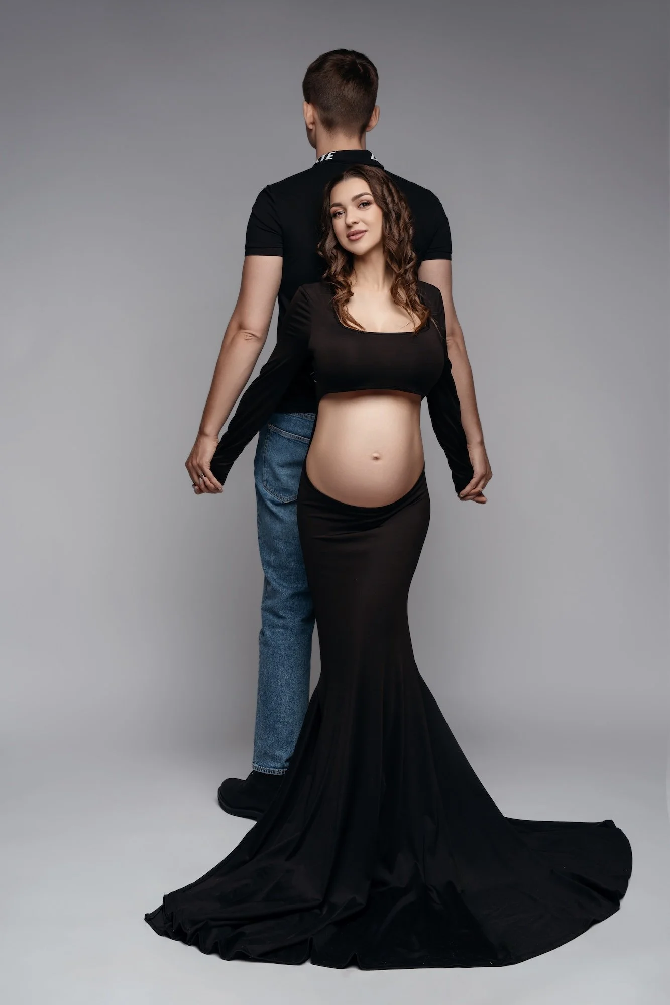  Elegant all-black couples maternity look: wife in a black gown with a long train, husband in black polo and jeans, Chicago studio grey backdrop 