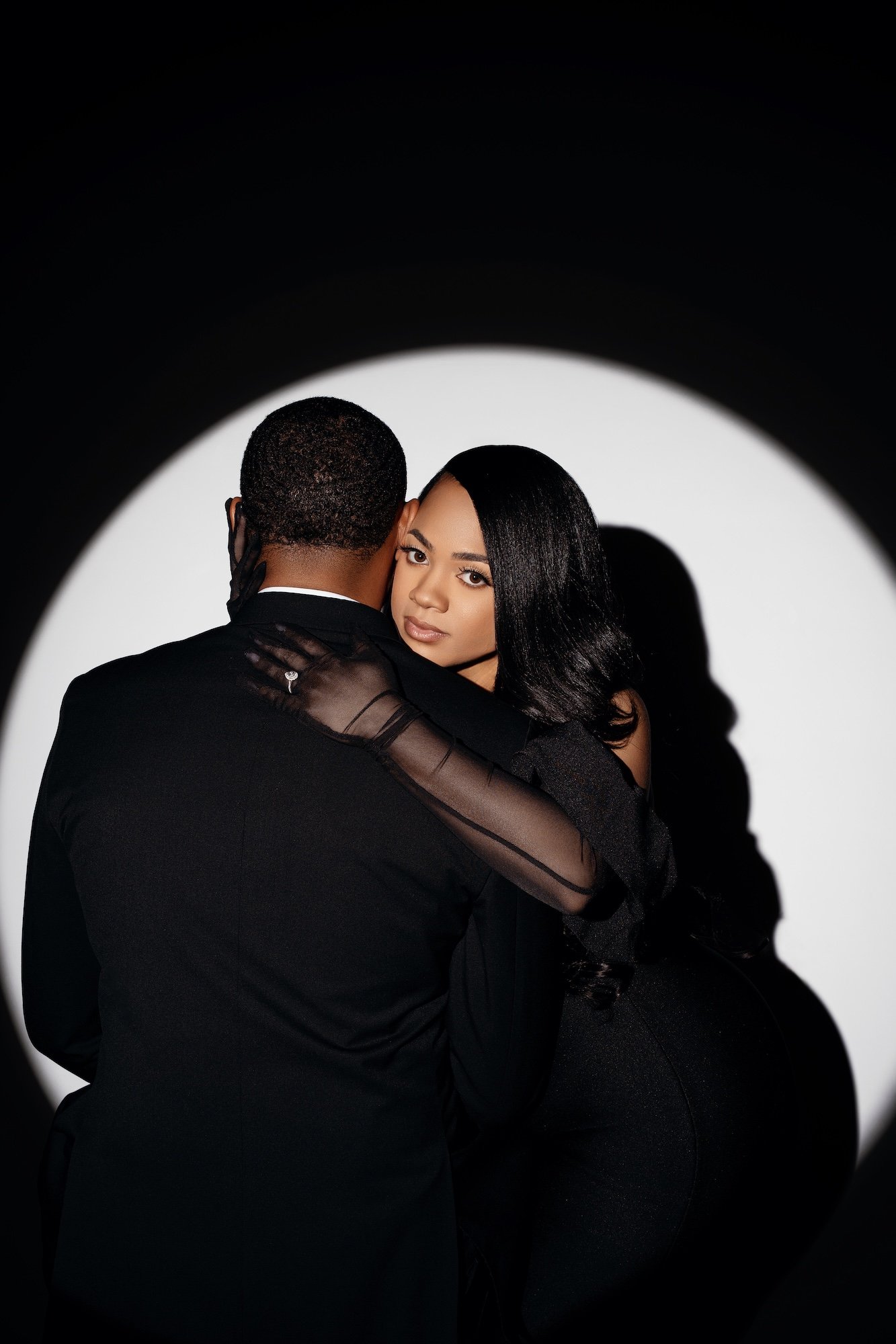 Dramatic spotlight engagement portrait, couple embracing in circle of light on black background, woman in black dress with sheer gloves, Chicago studio