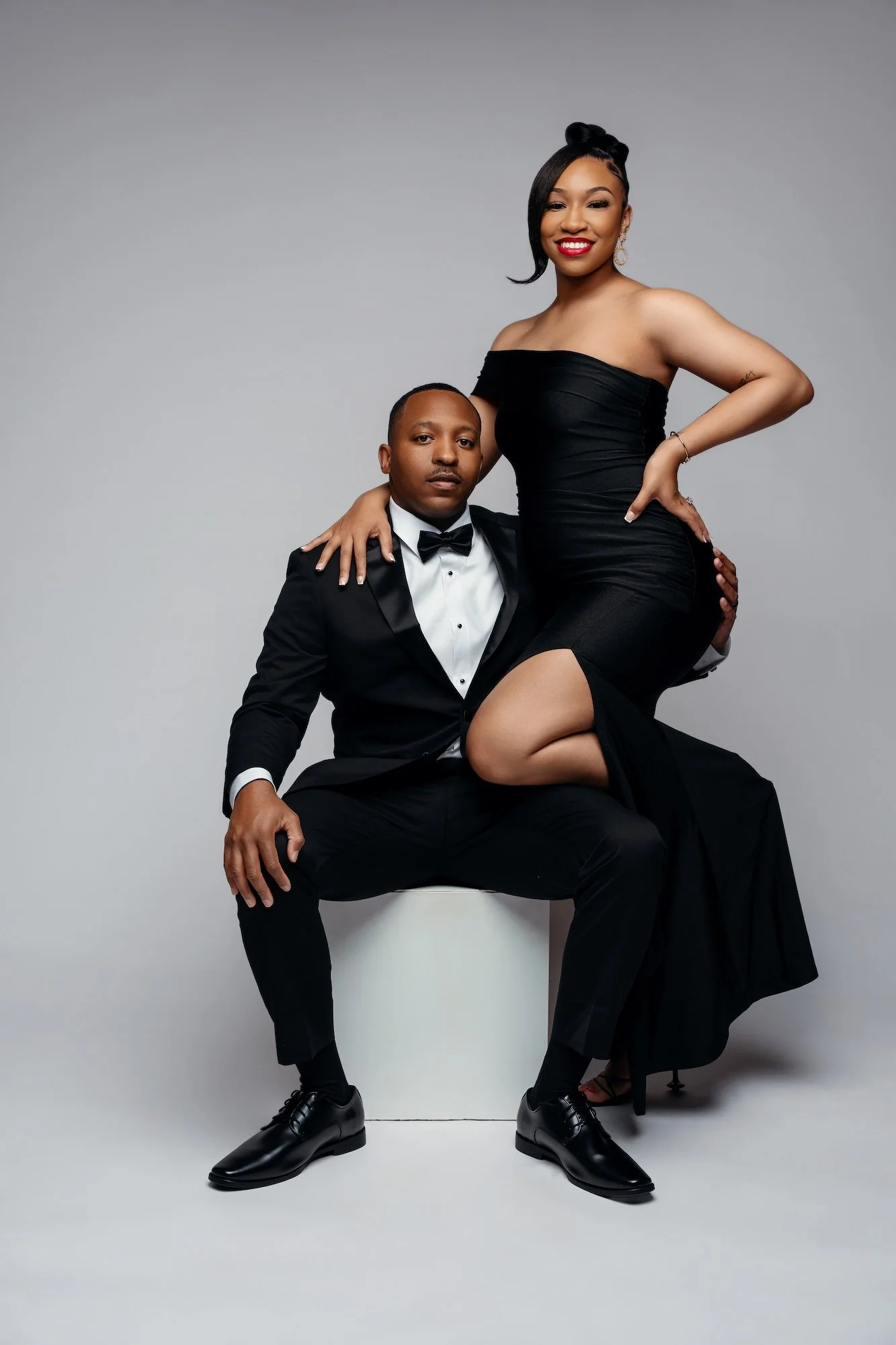 Elegant engagement portrait, woman in black off-shoulder dress sitting on man's lap in tuxedo, formal studio photography Chicago