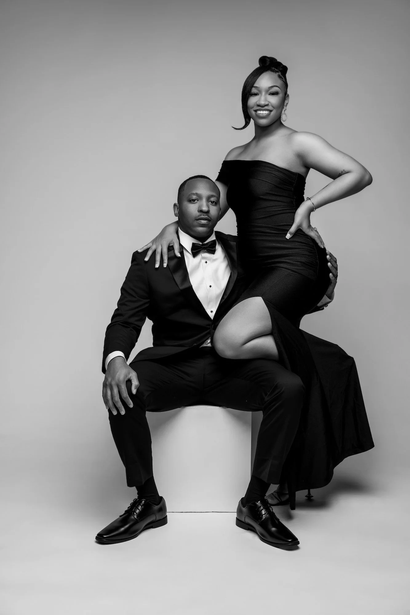 Black and white elegant engagement photo, woman in black dress on man's lap in tuxedo, sophisticated studio portrait at Filaneyka