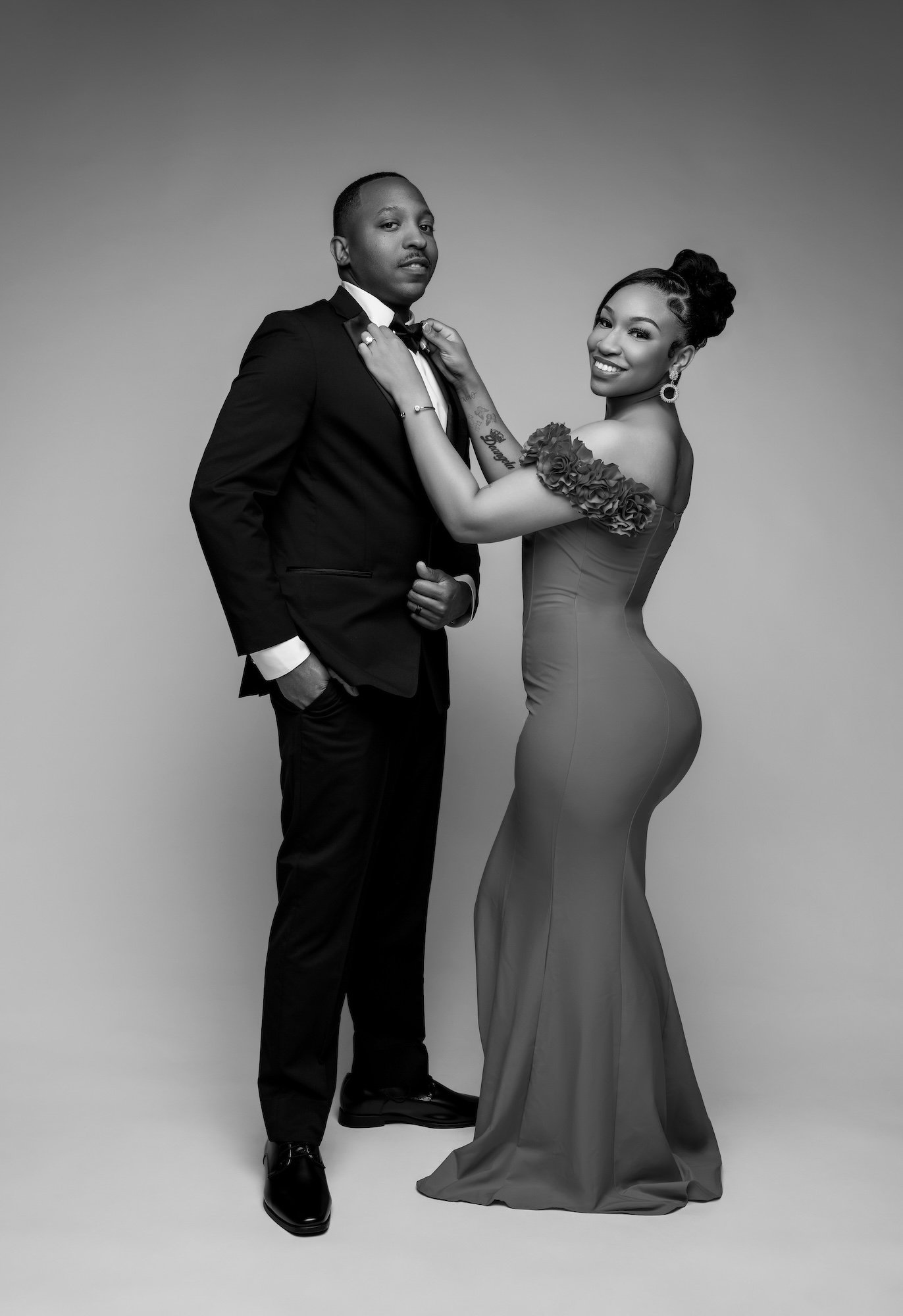 Black and white candid engagement photo, woman adjusting man's bowtie in red dress, playful studio moment at Filaneyka