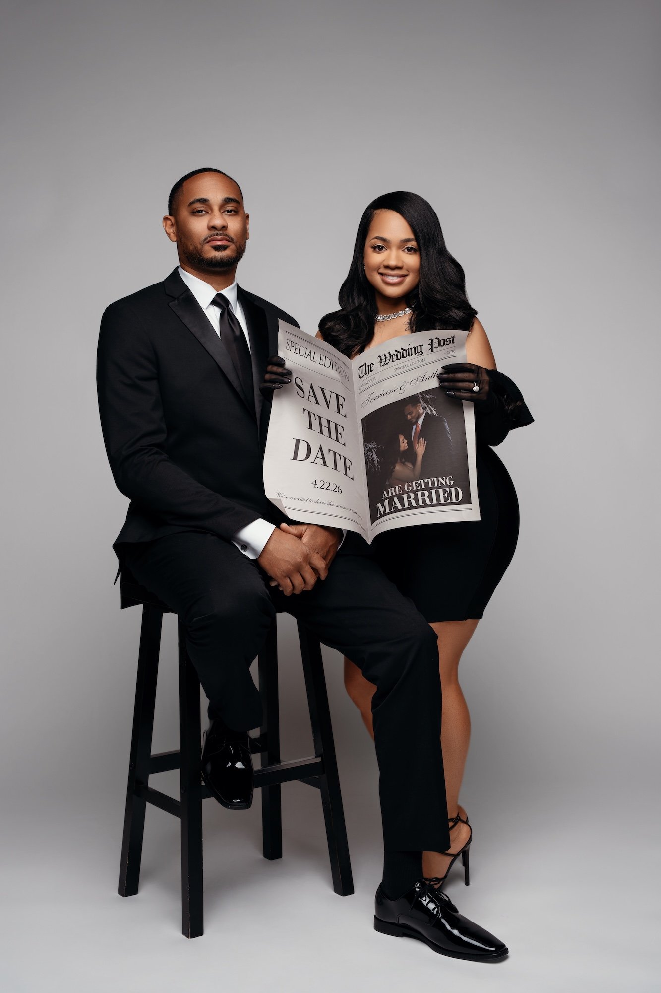 Save the date engagement portrait, couple sitting on stool holding "The Wedding Post" newspaper, black outfits, gray backdrop, Chicago photographer
