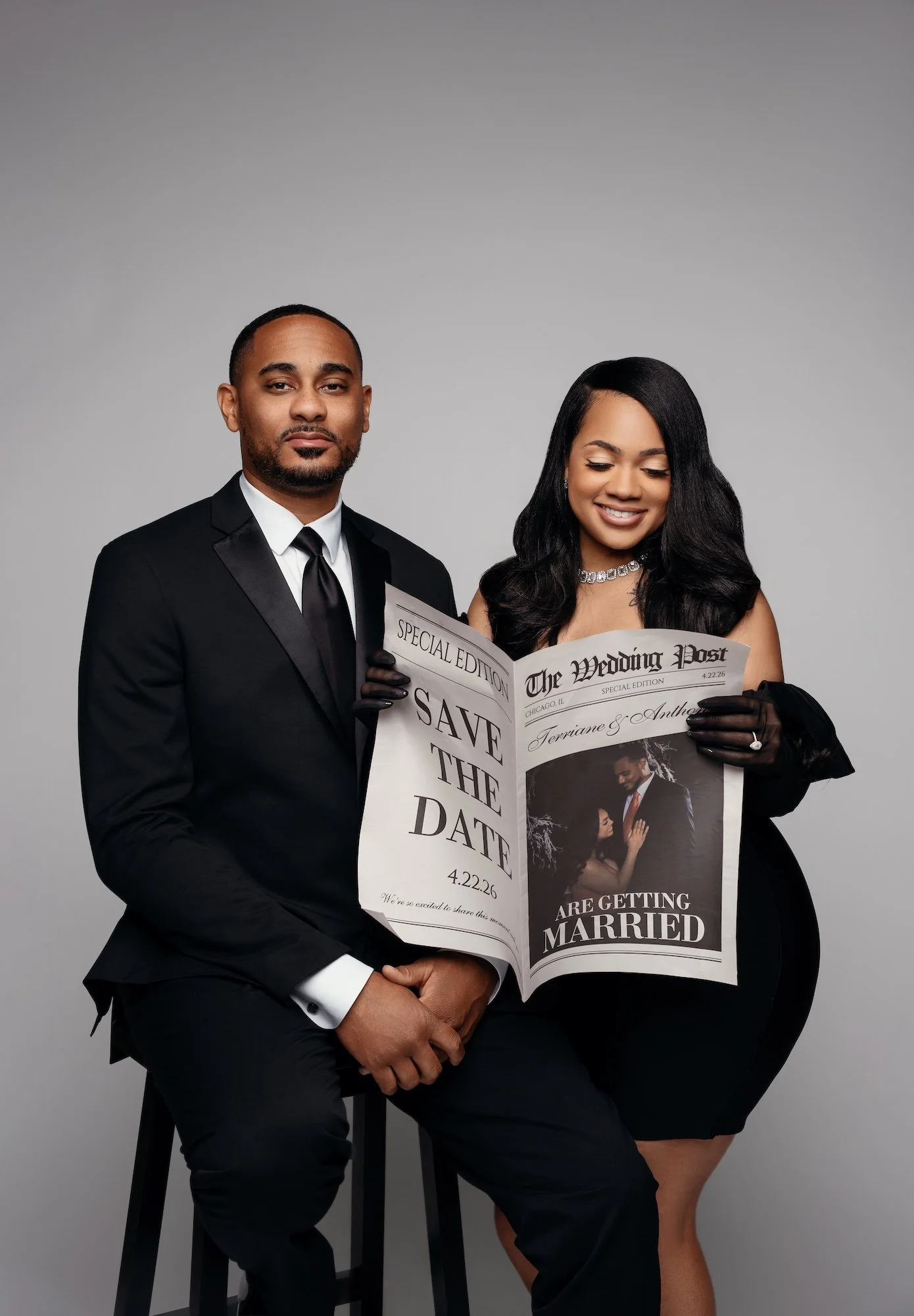Save the date engagement photo, couple holding "The Wedding Post" newspaper with save the date 4.22.26, woman in black dress, studio portrait Chicago