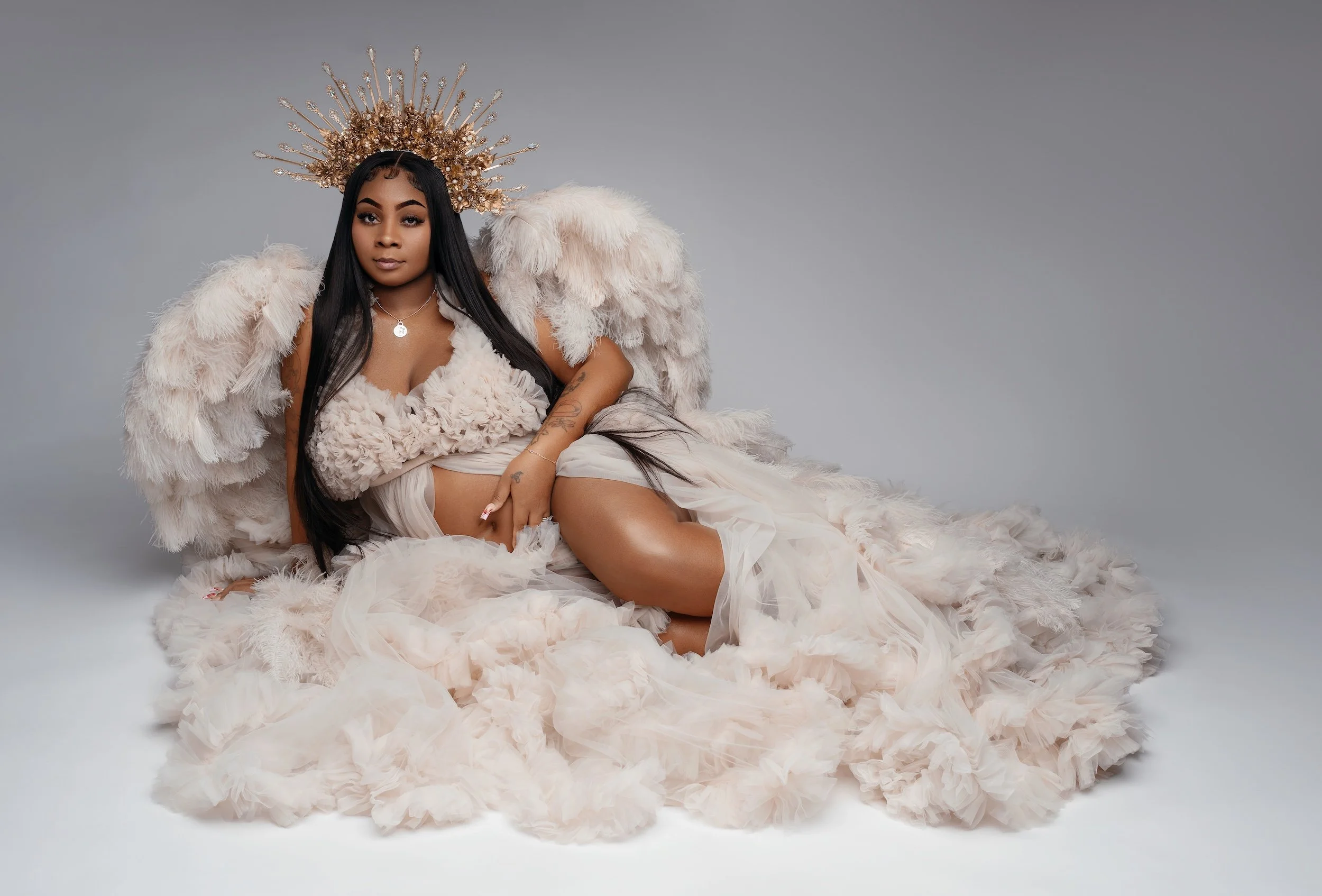 Stunning curvy woman as modern angel with golden spiked crown, oversized cream feather wings, long black hair and sheer beige tulle dress in elegant studio portrait