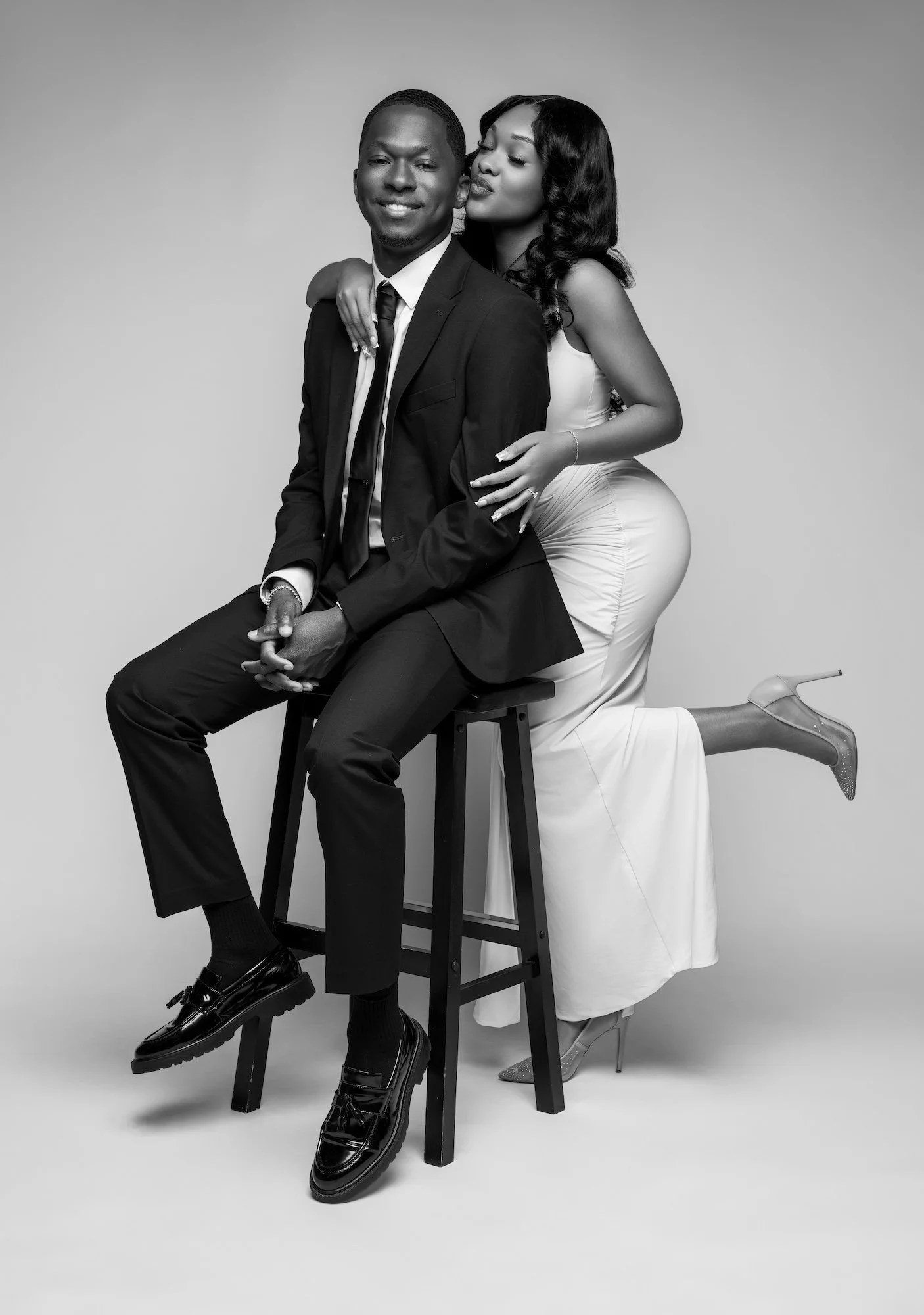 Black and white candid engagement photo, woman kissing man's cheek on stool, joyful studio moment at Filaneyka Photo Studio