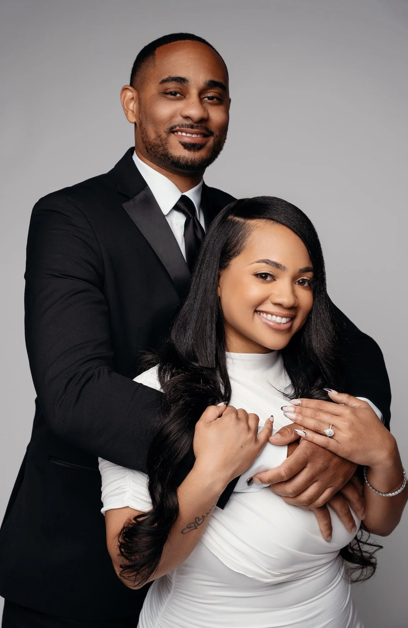 Warm engagement portrait, man in black suit hugging woman in white turtleneck from behind, both smiling, gray backdrop, Chicago studio photographer