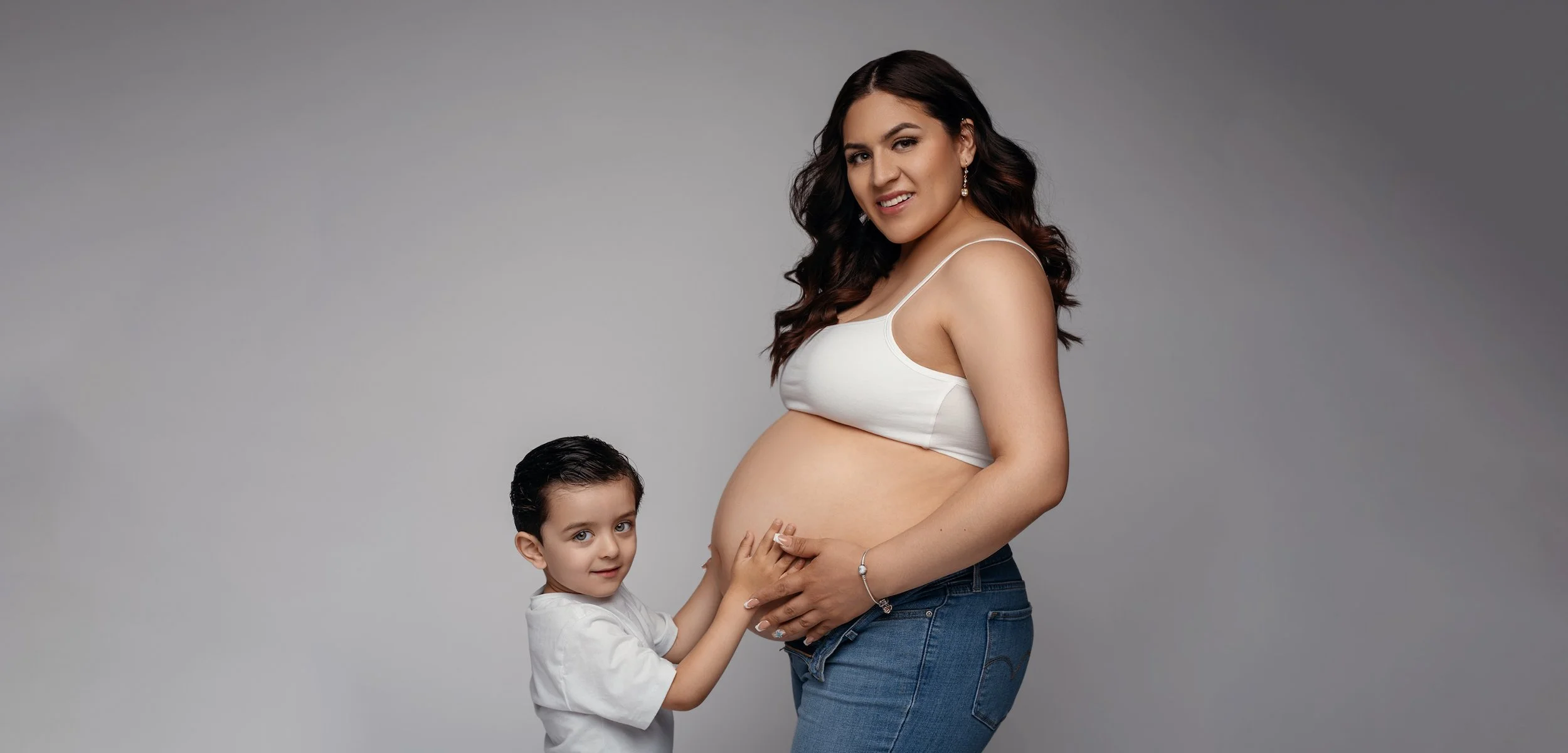 Pregnant mother in white crop top embracing baby bump with her young son, grey studio background