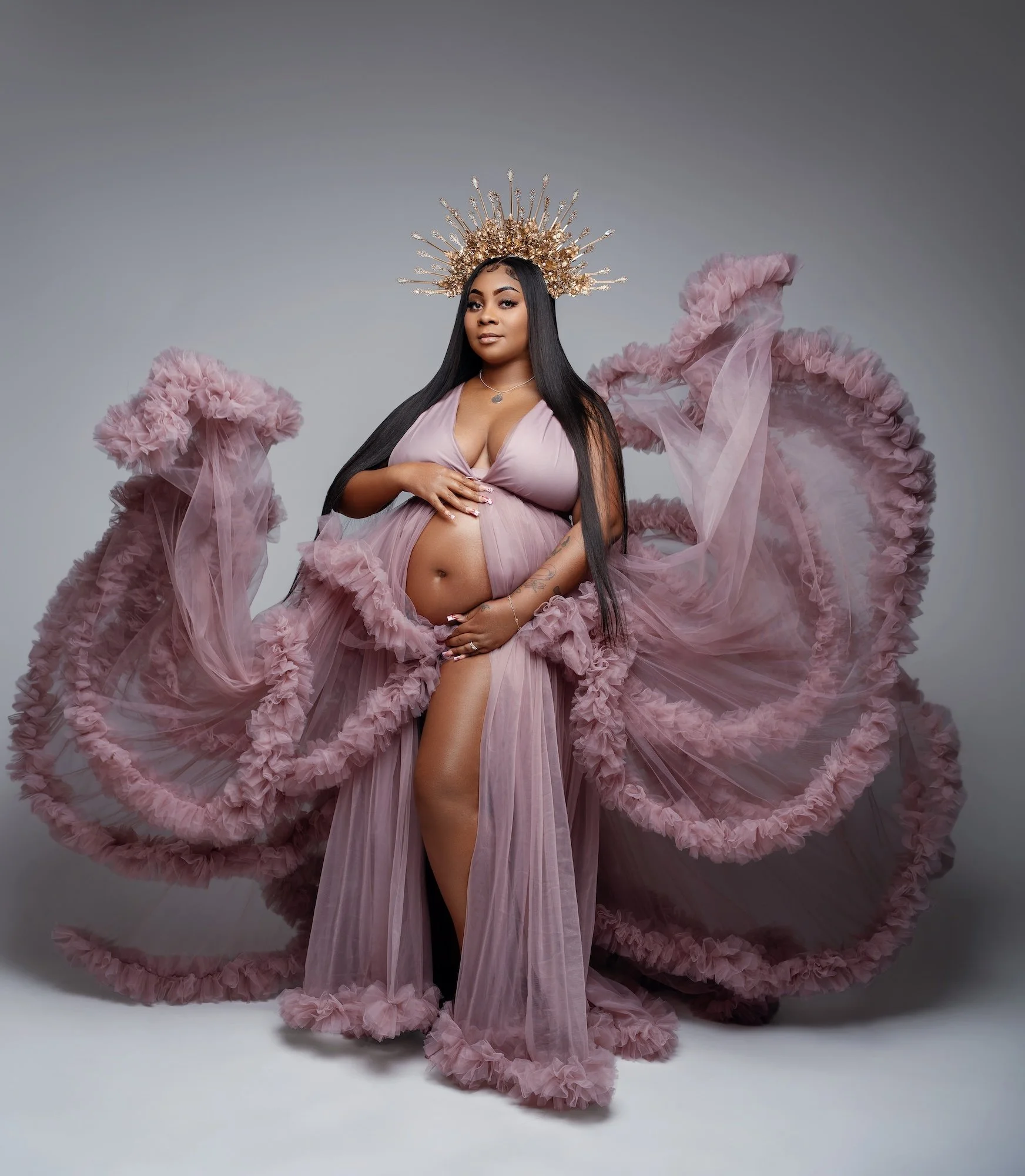 Elegant studio maternity portrait of a pregnant woman with long straight black hair wearing a dramatic flowing purple ruffled tulle gown and an ornate golden crown. She poses confidently with both hands gently placed on her bare pregnant belly agains