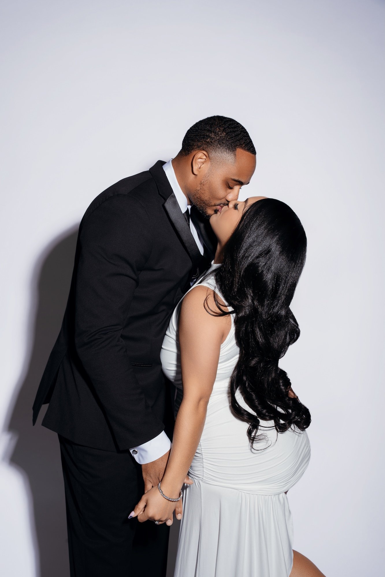 Romantic engagement kiss, man in black suit kissing woman in white dress against white backdrop, dramatic shadow, studio photography Chicago