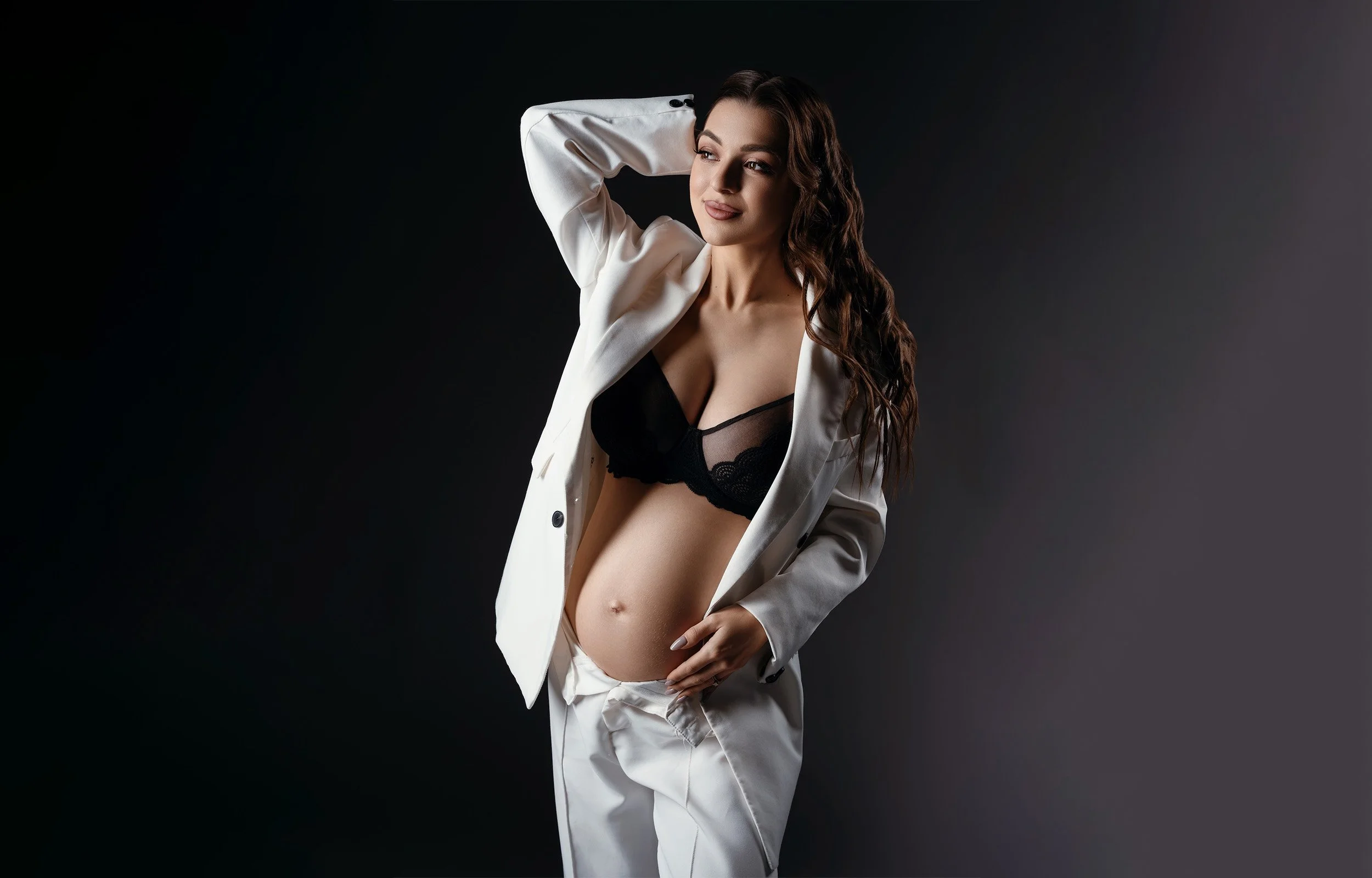 Stylish maternity portrait of a pregnant woman wearing an open white suit over black lace lingerie, posing confidently with one hand behind her head and the other on her baby bump against a dark studio backdrop.