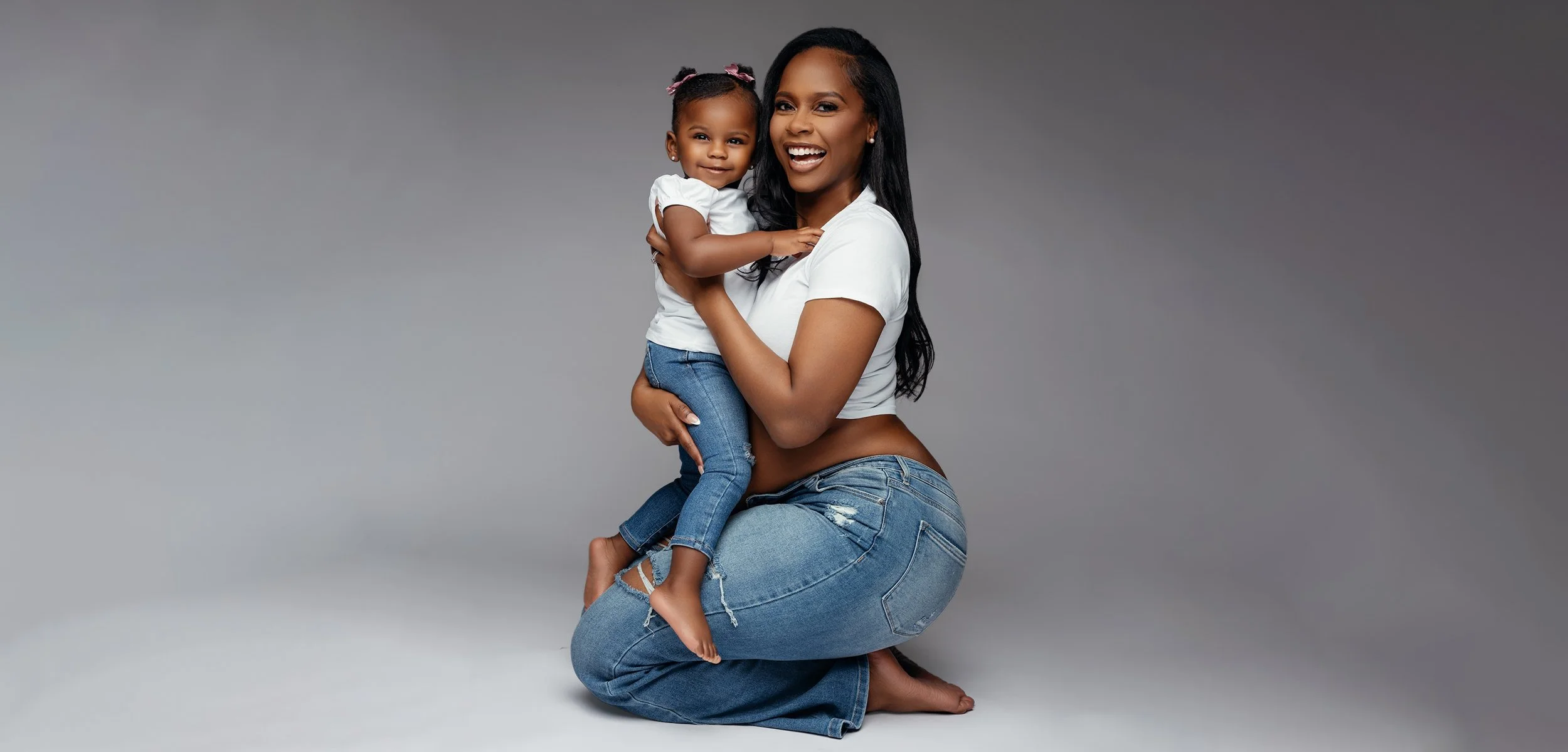 Wide studio portrait of joyful pregnant mother holding her toddler daughter against soft gray background