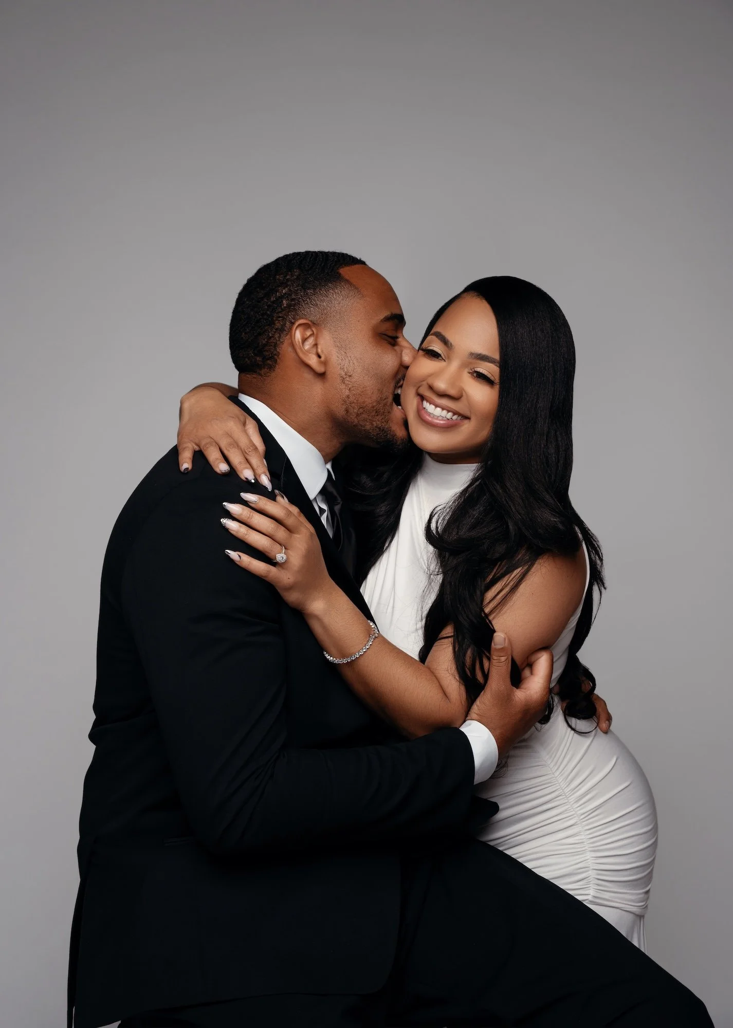 Joyful engagement photo, man kissing smiling woman's cheek while embracing, gray backdrop, candid studio portrait Chicago